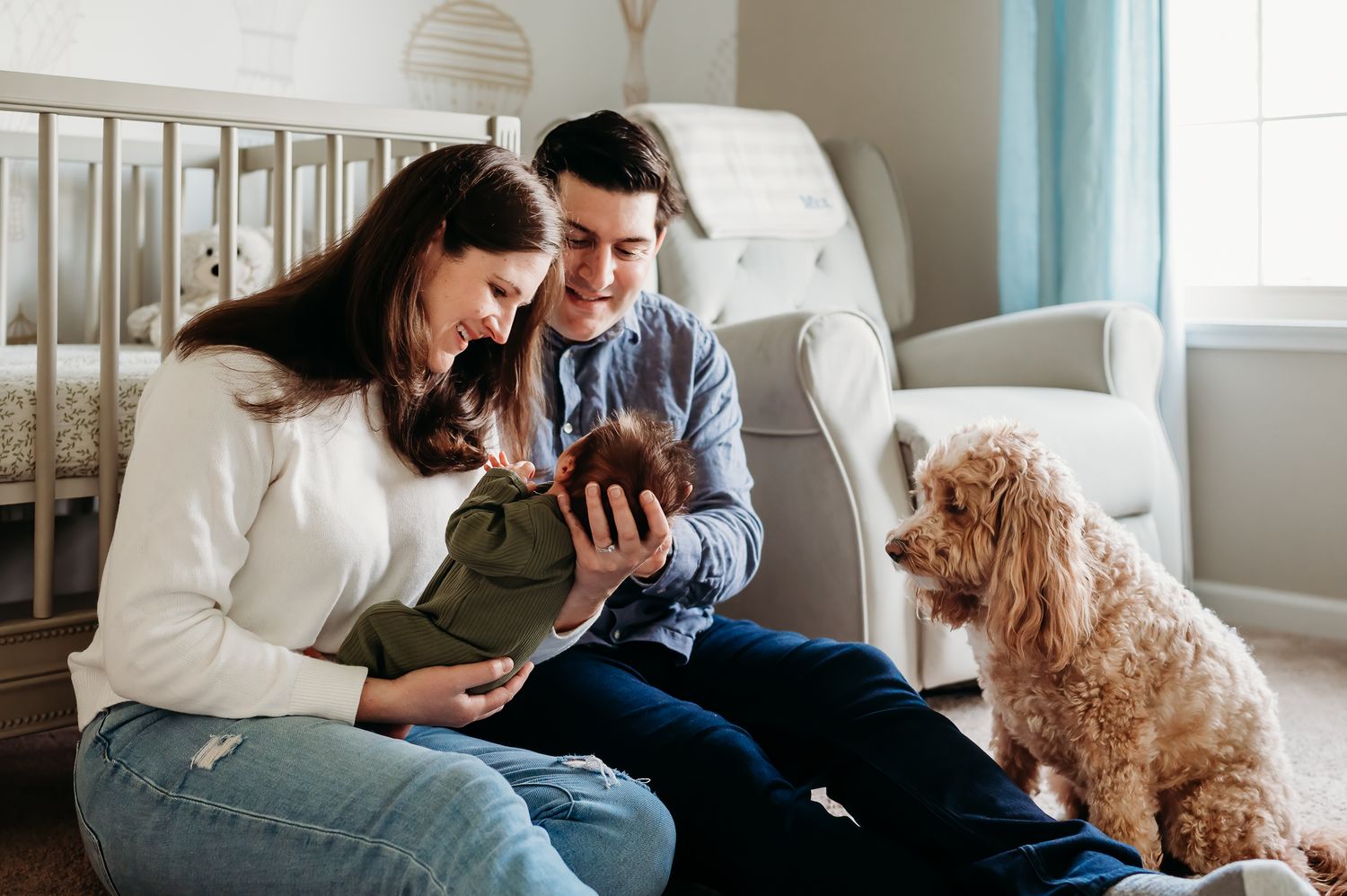 A couple sits together by crib with a baby and their goldendoodle dog in a bright nursery room in Noblesville, Indiana.