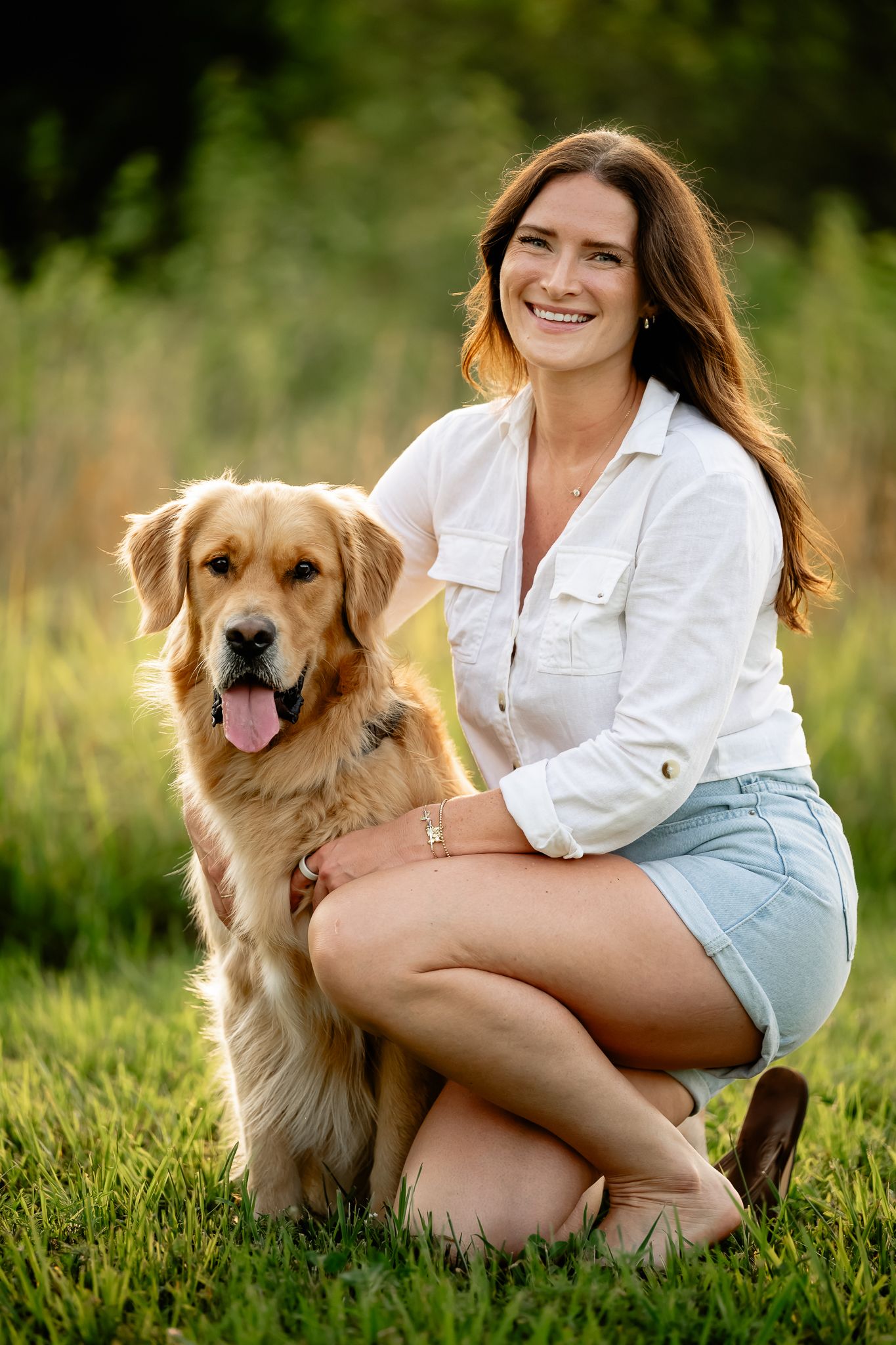 Smiling young woman in white shirt and denim shorts kneeling with a golden retriever on green grass during dog photoshoot in Plainfield, IN.