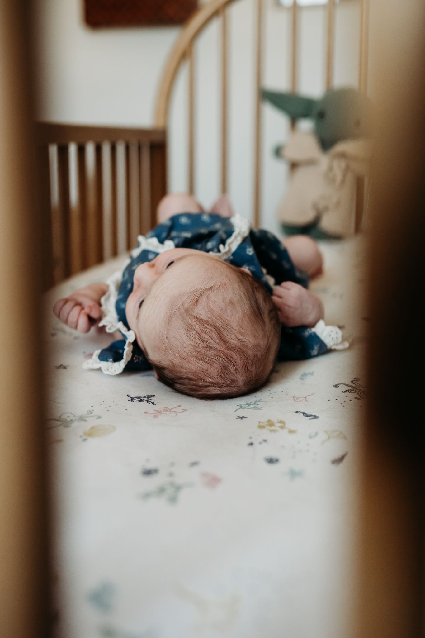 Baby sleeping peacefully in a crib wearing blue clothing on a patterned sheet, viewed through crib slats.