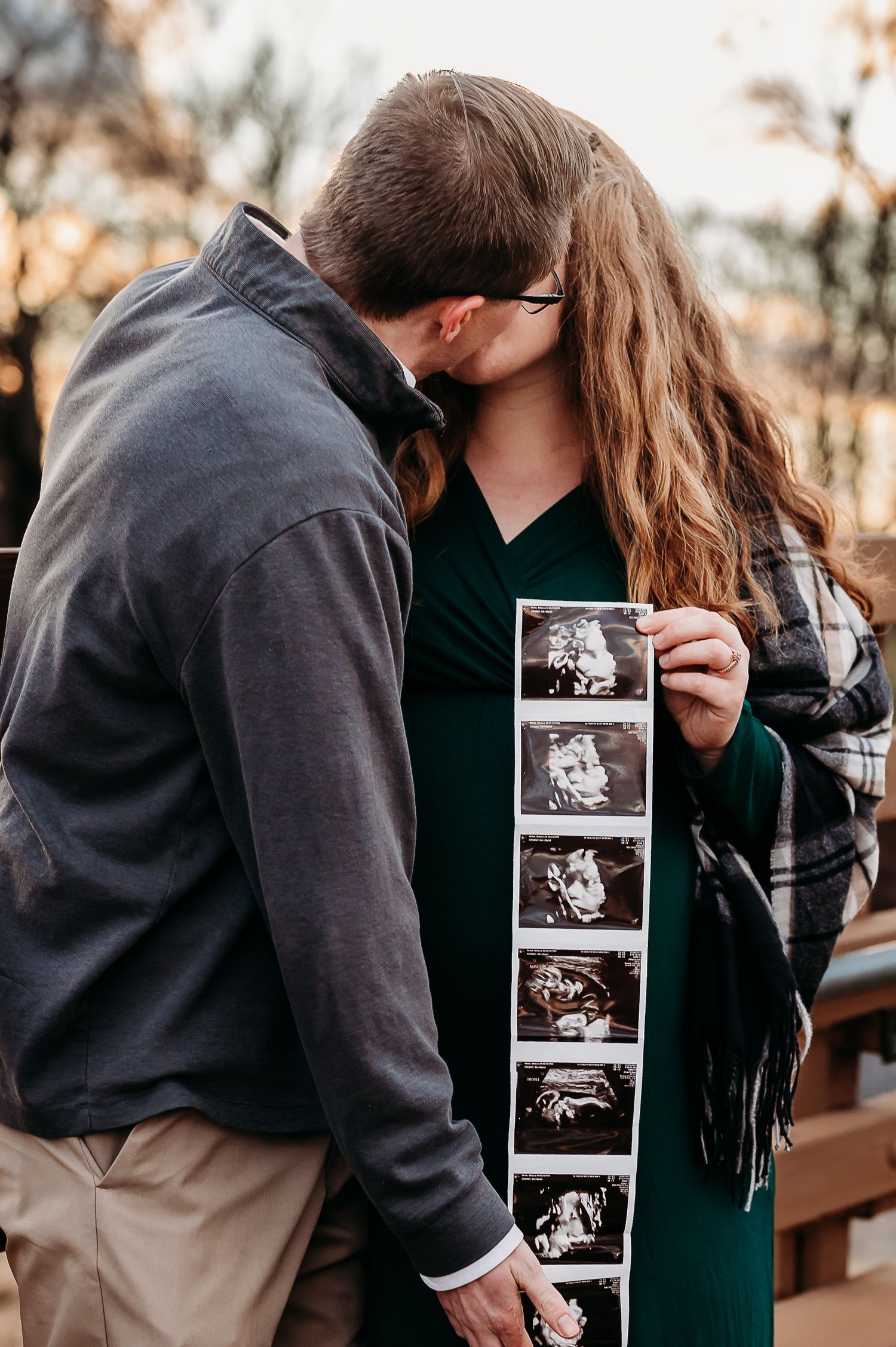 Couple kissing while holding ultrasound photos, announcing pregnancy outdoors during sunset maternity photoshoot in Plainfield, Indiana.
