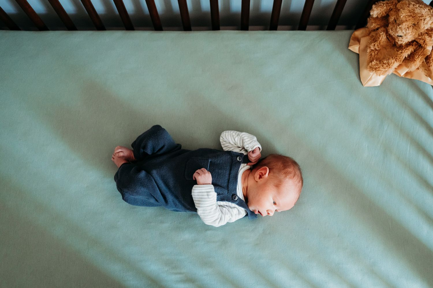 Small baby wearing navy blue outfit lies on teal mattress sheet in Indiana nursery during photoshoot with Sara House Photography.