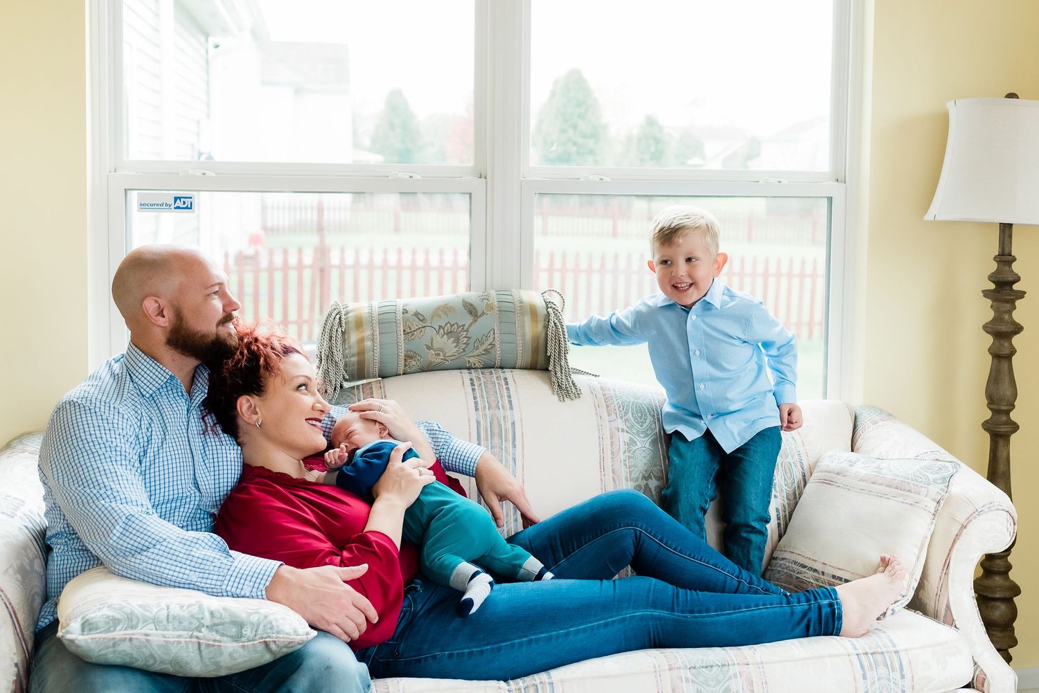 Parents relax with baby boy and big brother on a light colored couch in a sunny living room by a window during newborn photos in Avon, IN.