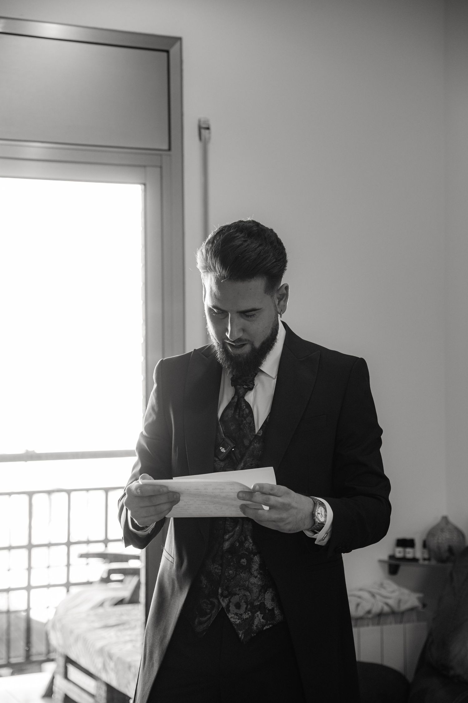 A person in a dark suit and tie reads documents while standing in a minimalist room with natural light from a window.