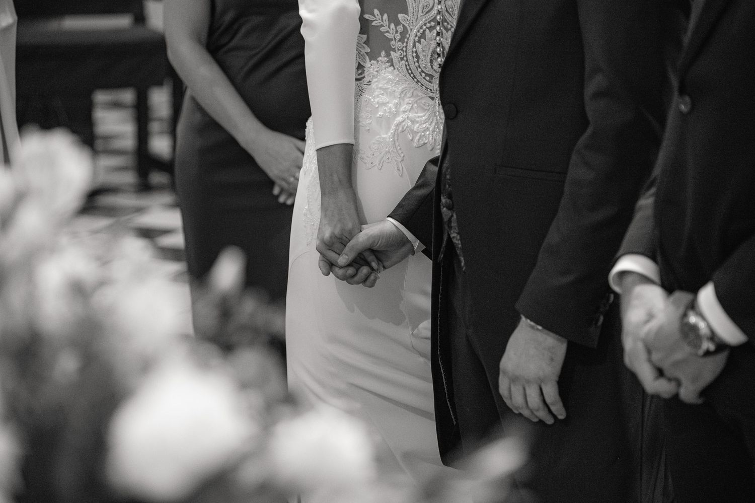 Black and white photo showing couple holding hands at wedding ceremony with bride wearing white lace dress.