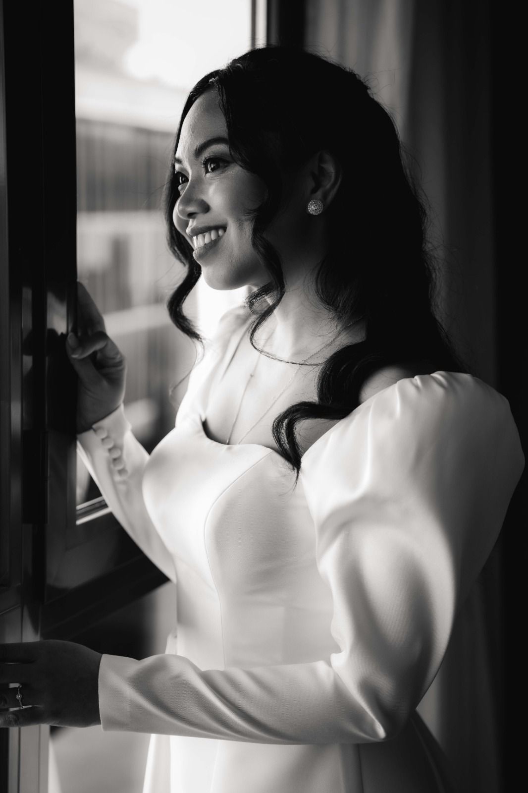 Black and white portrait of someone in a white dress with puff sleeves smiling by a window in natural light.