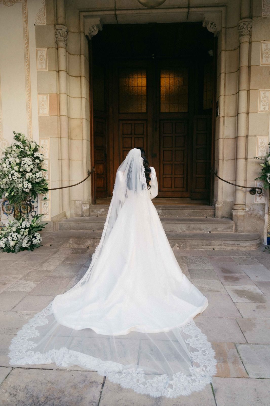 A bride in a flowing white wedding gown and long lace veil stands before ornate wooden church doors.