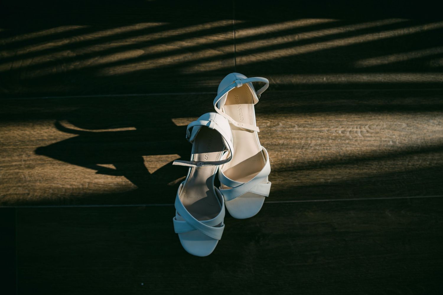 Elegant white bridal sandals with ankle straps cast dramatic shadows on a dark wooden surface in natural light.