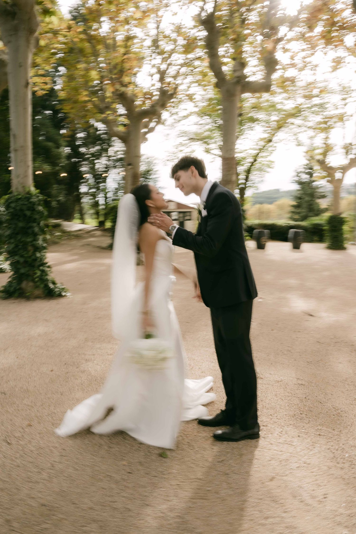 A couple in wedding attire dance and twirl together on a tree-lined path during their outdoor photo session.