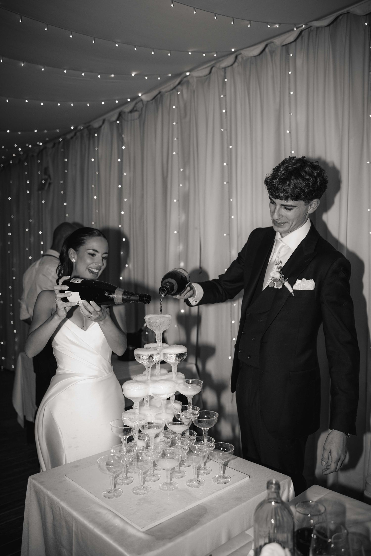 A series of black and white photos showing a couple pouring champagne into a tower of glasses at their wedding reception.