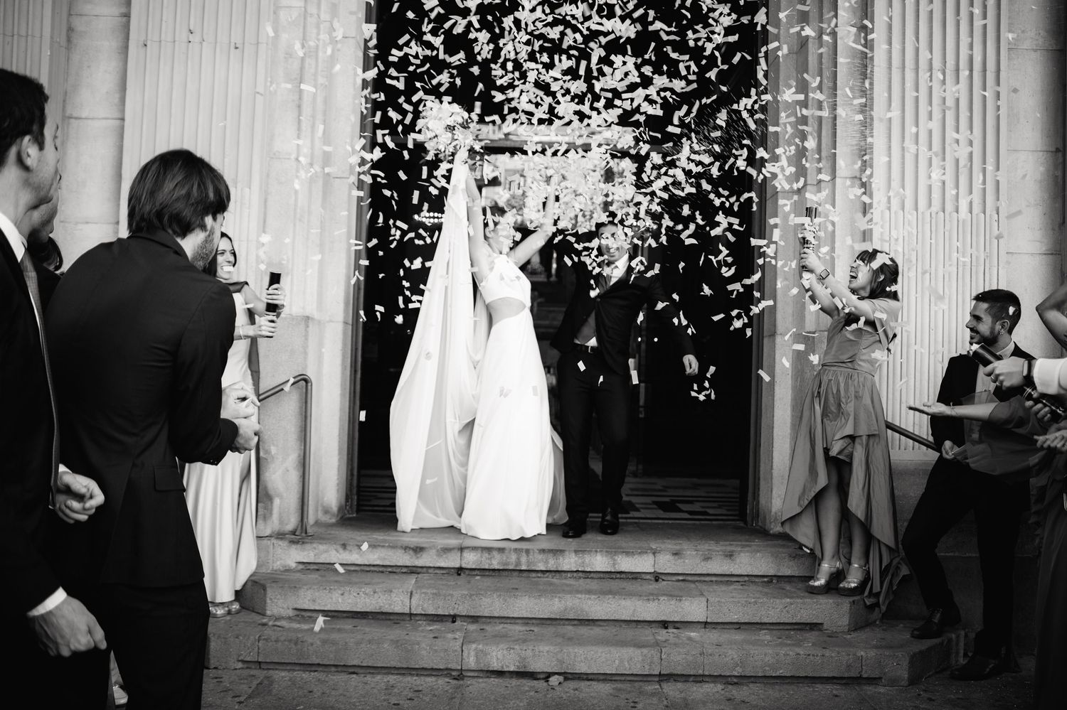 Wedding guests celebrate with confetti and flower petals as newlyweds exit the ceremony at the church steps.