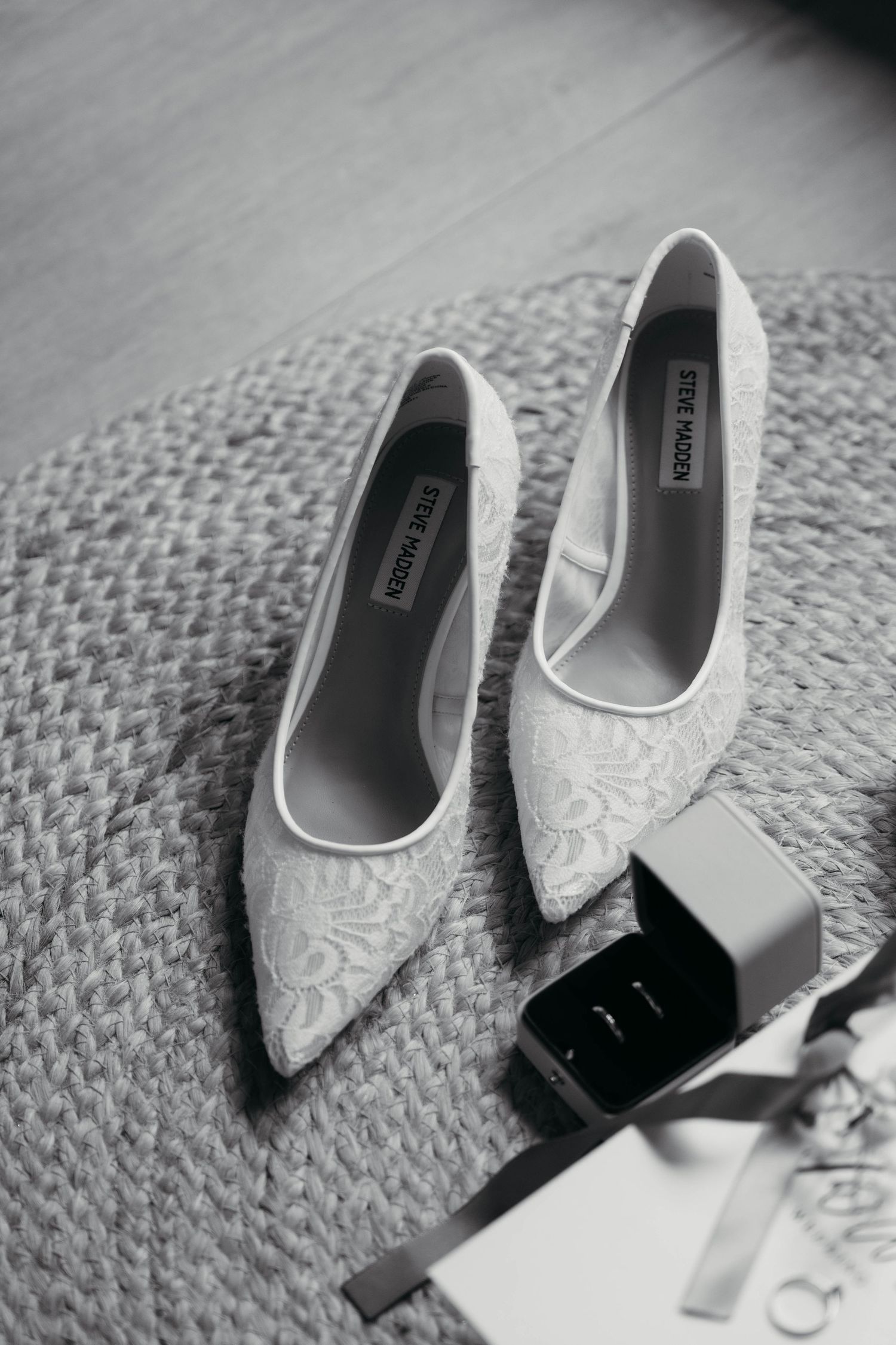 Black and white photo of Steve Madden lace bridal heels beside a ring box and wedding rings on a woven rug.