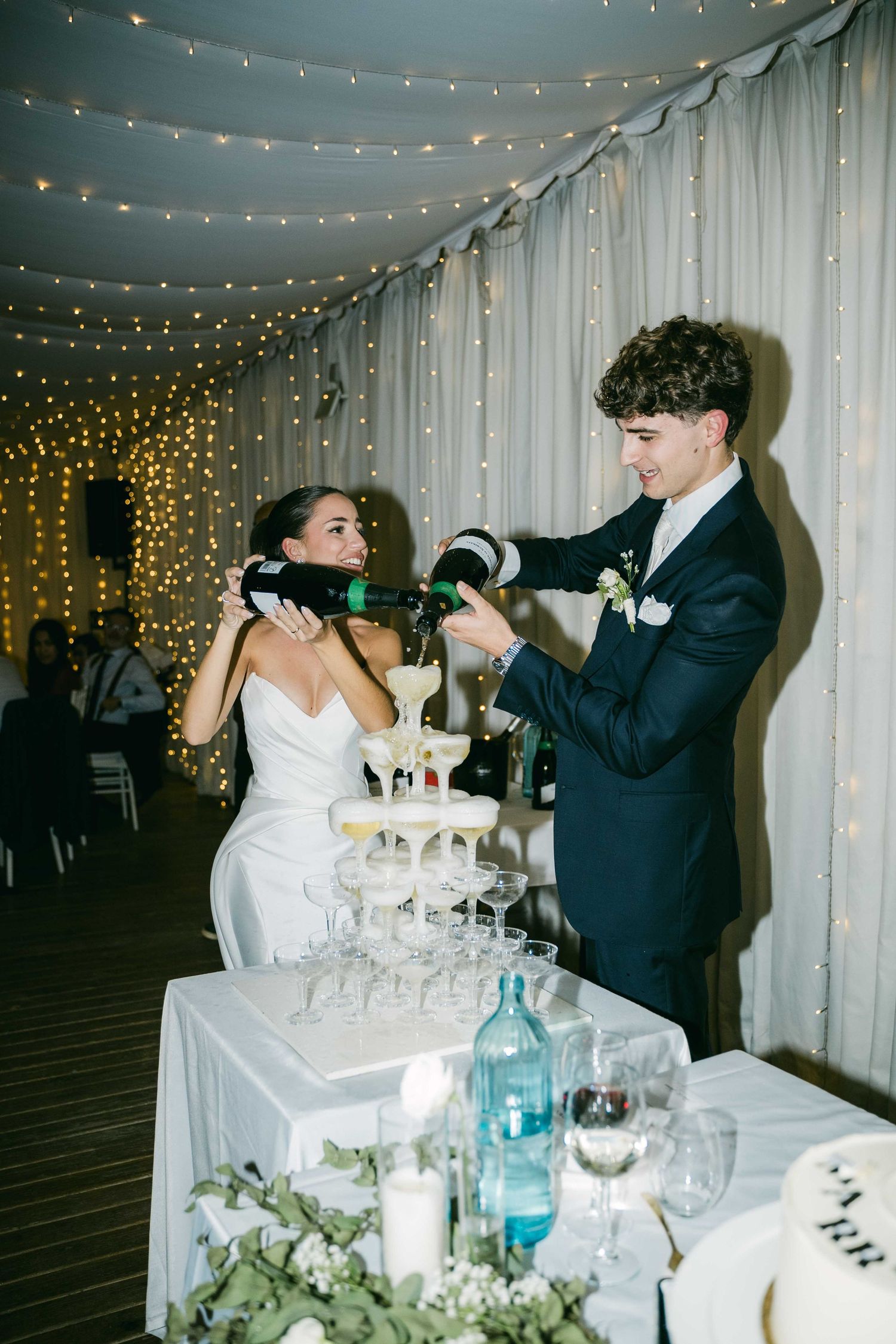 Newlyweds playfully feed each other champagne at their wedding reception surrounded by twinkling lights and white decor.