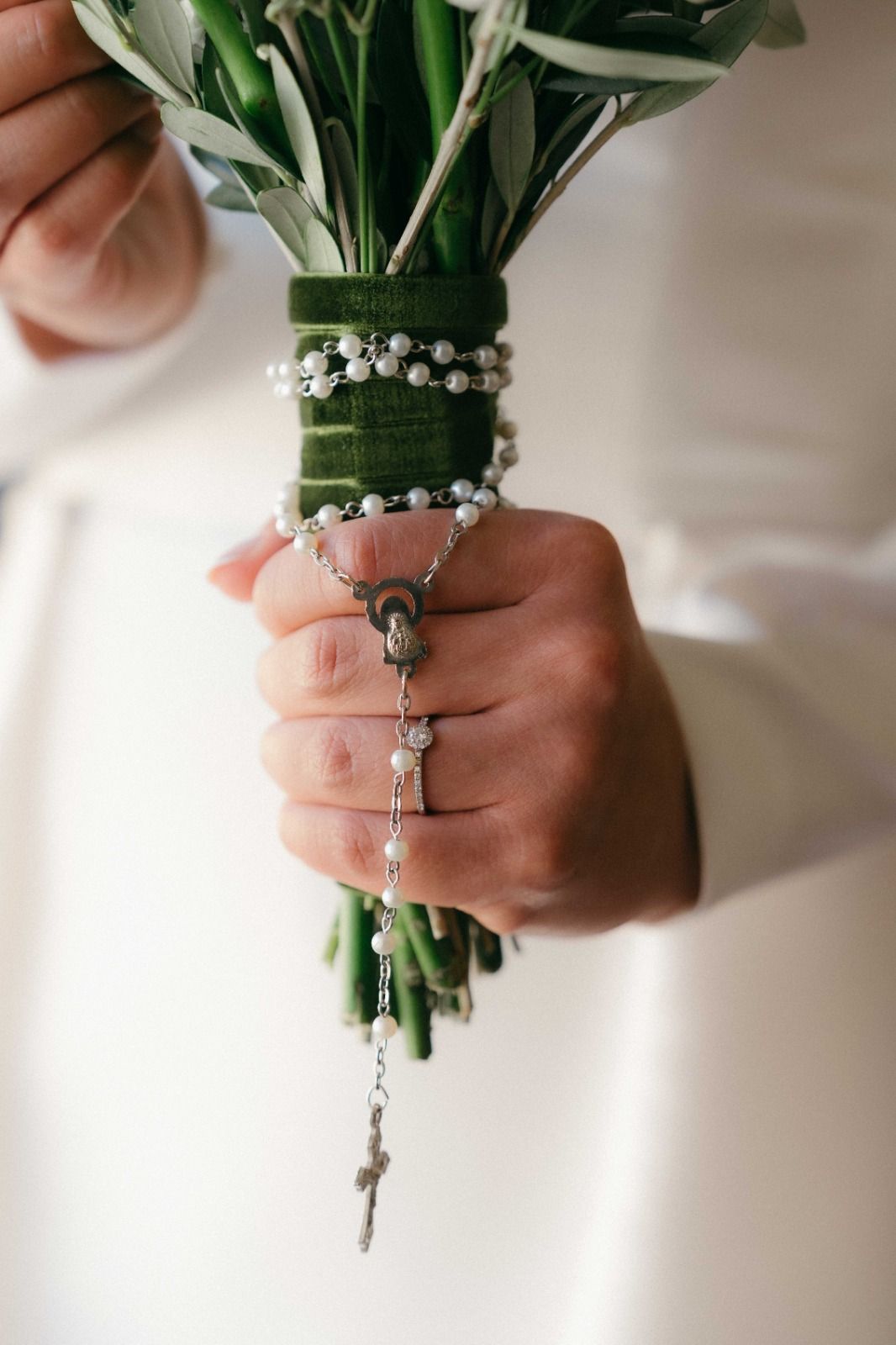 Close-up view of hands holding a bridal bouquet wrapped with white lily of the valley flowers and green stems.