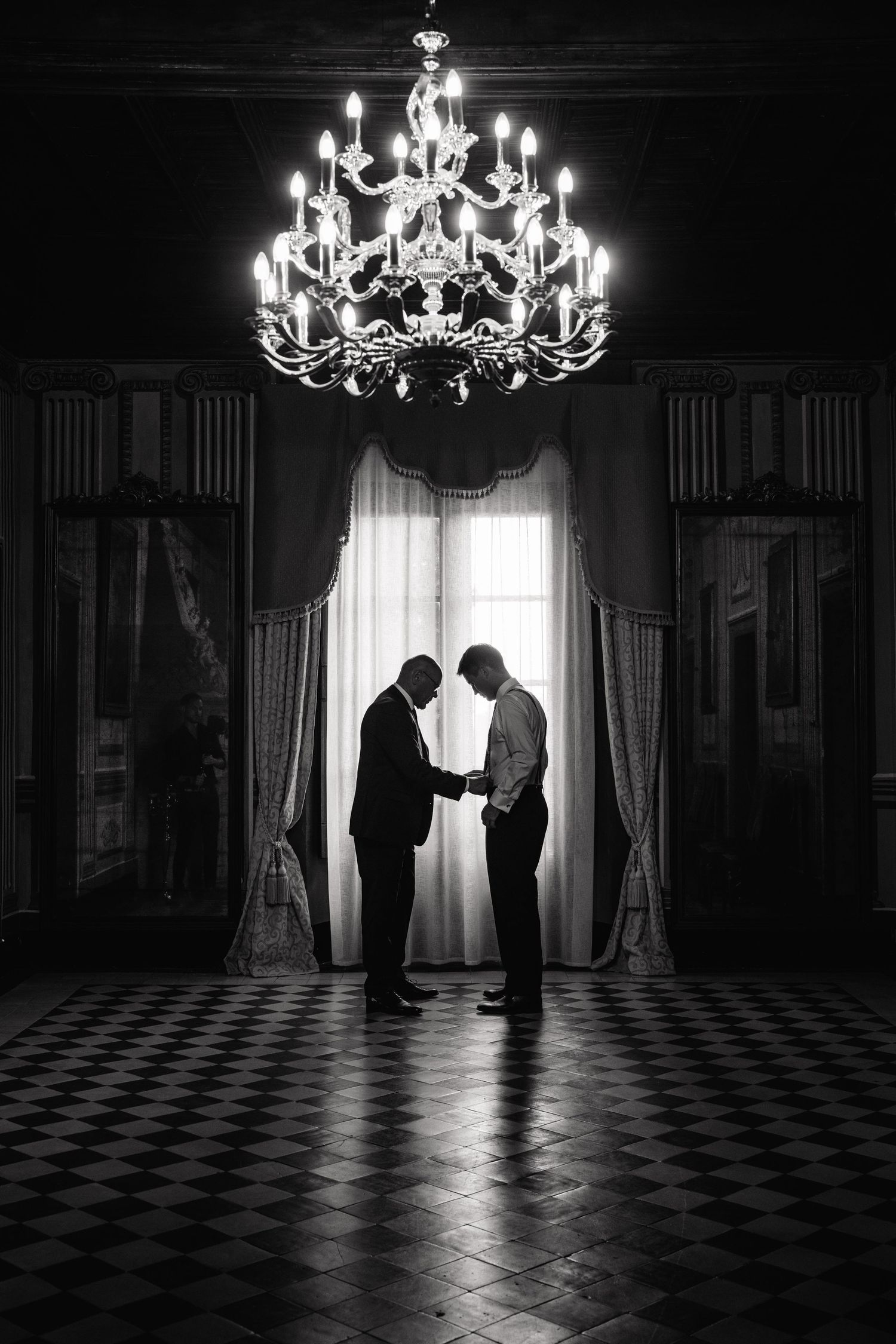 A dramatic silhouette of two figures beneath an ornate crystal chandelier in a grand ballroom.