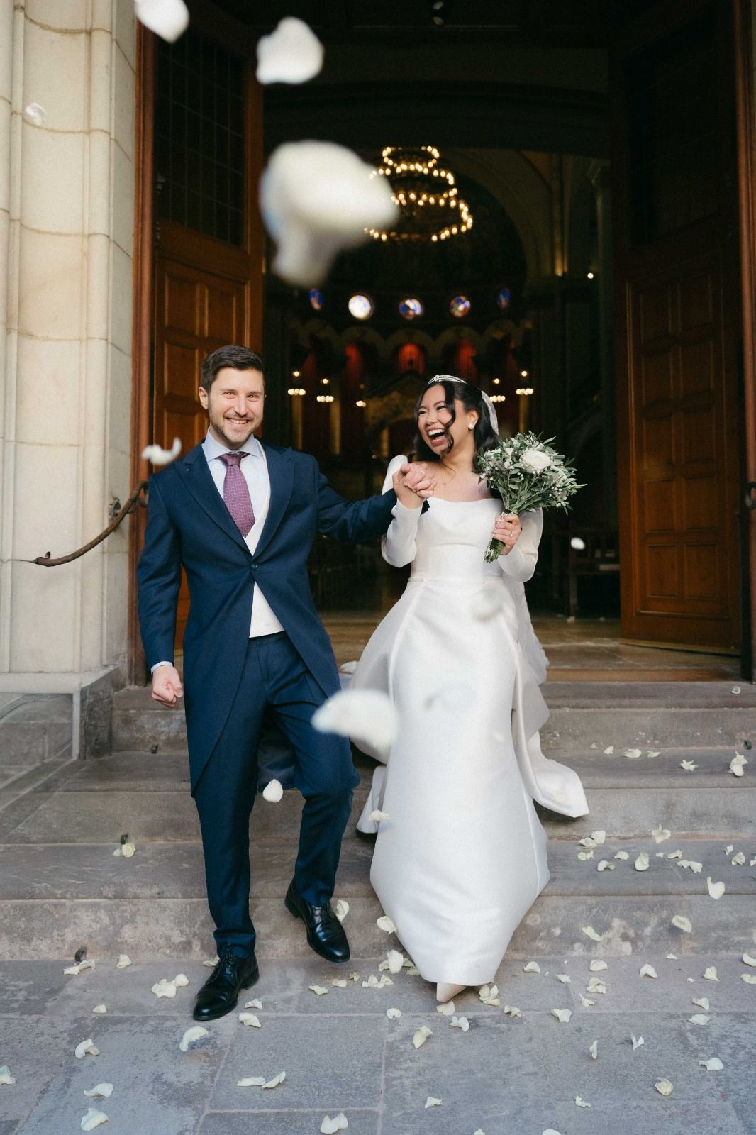 A joyful wedding couple exits their ceremony venue while white flower petals fall around them.