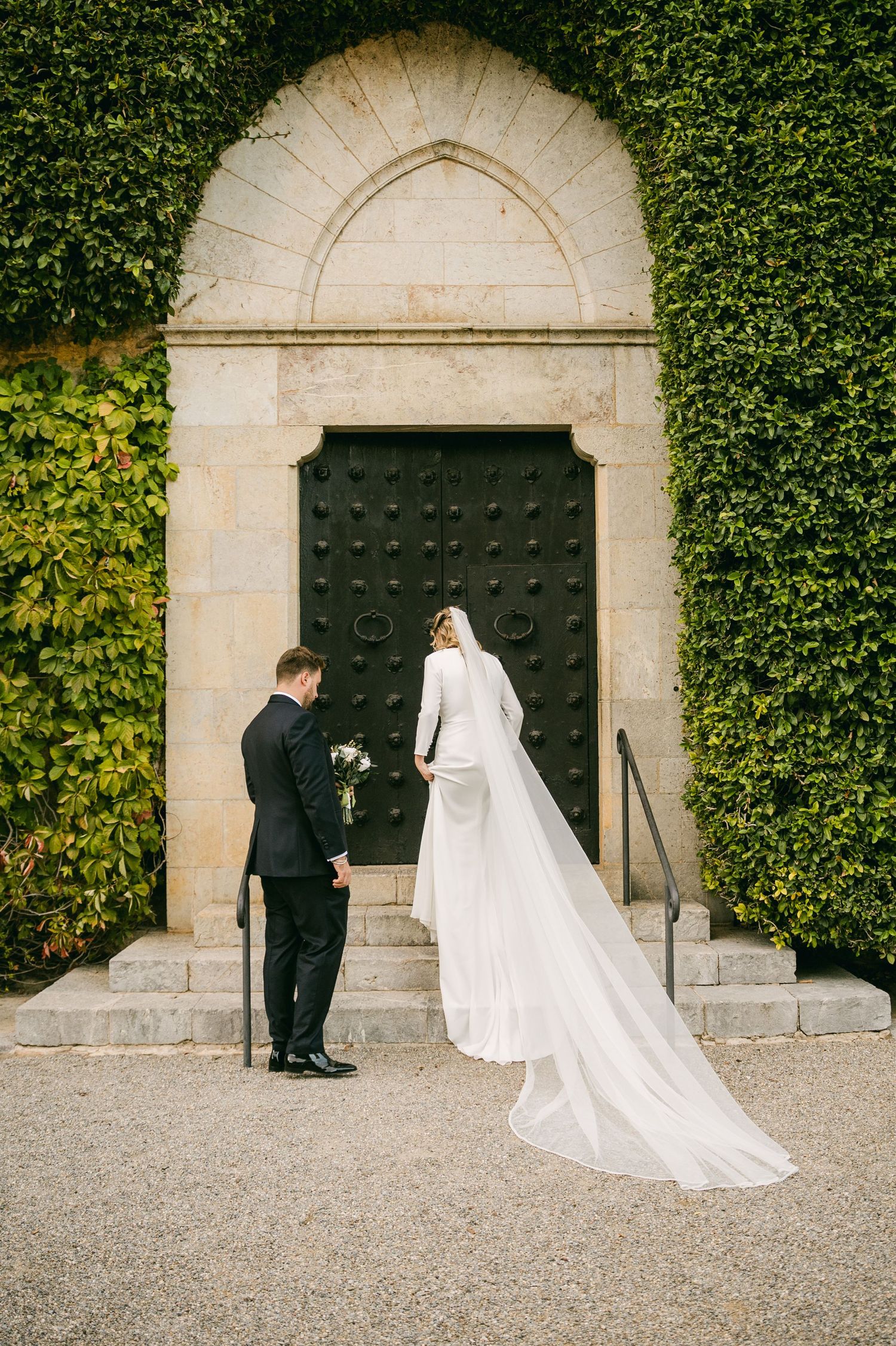 A couple stands before a grand ivy-covered archway with a large black door.