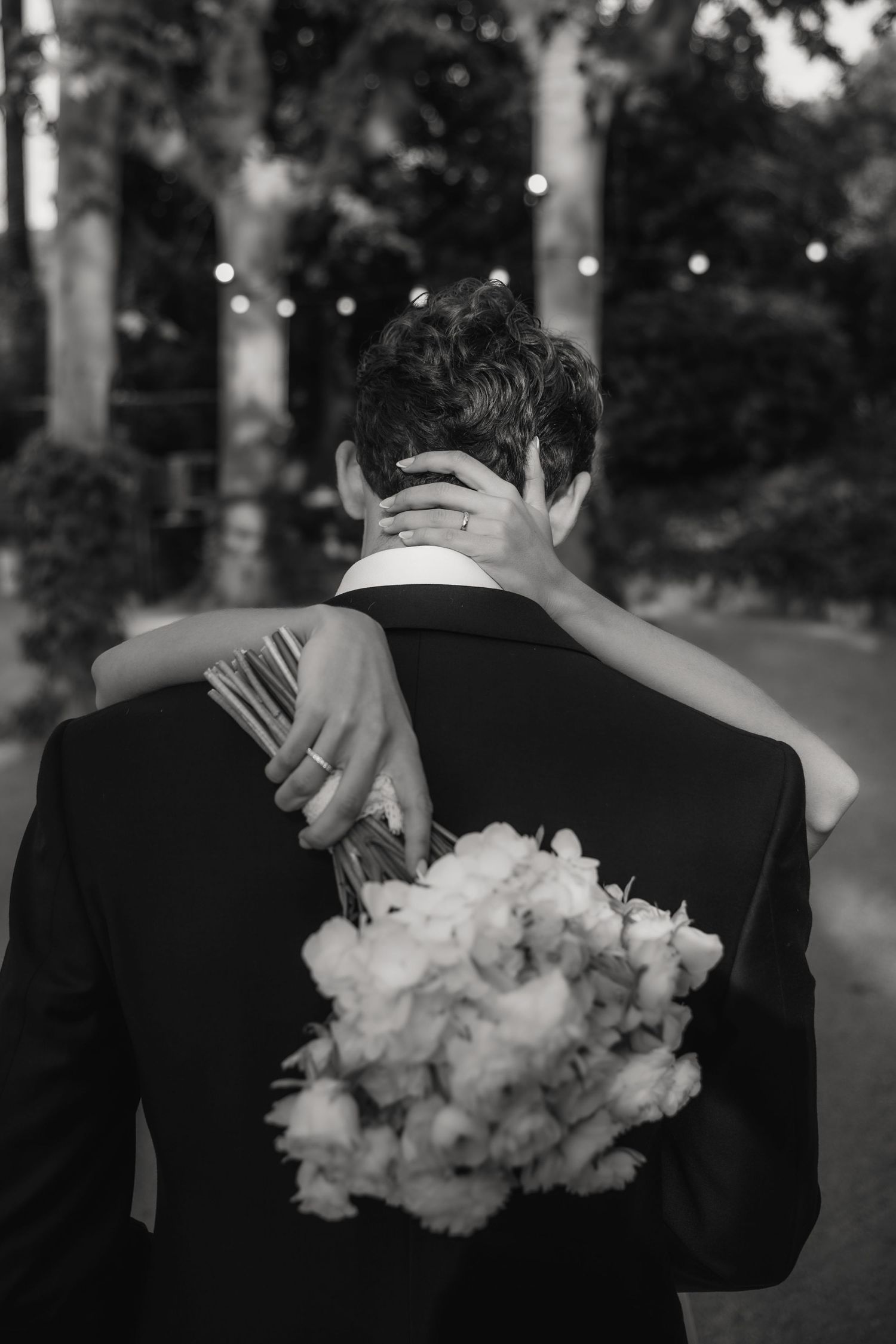 Black and white romantic wedding photo showing couple embracing with bride's hands on groom's neck and bouquet visible.
