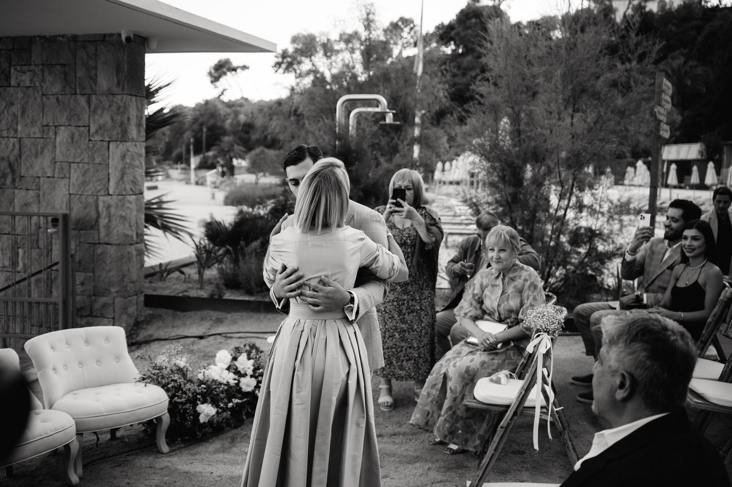 A black and white photograph of guests mingling on an outdoor patio with stone walls and furniture.