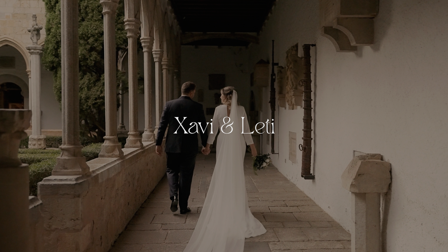 A couple walks down an elegant stone colonnade hallway with arches and pillars during their wedding celebration.