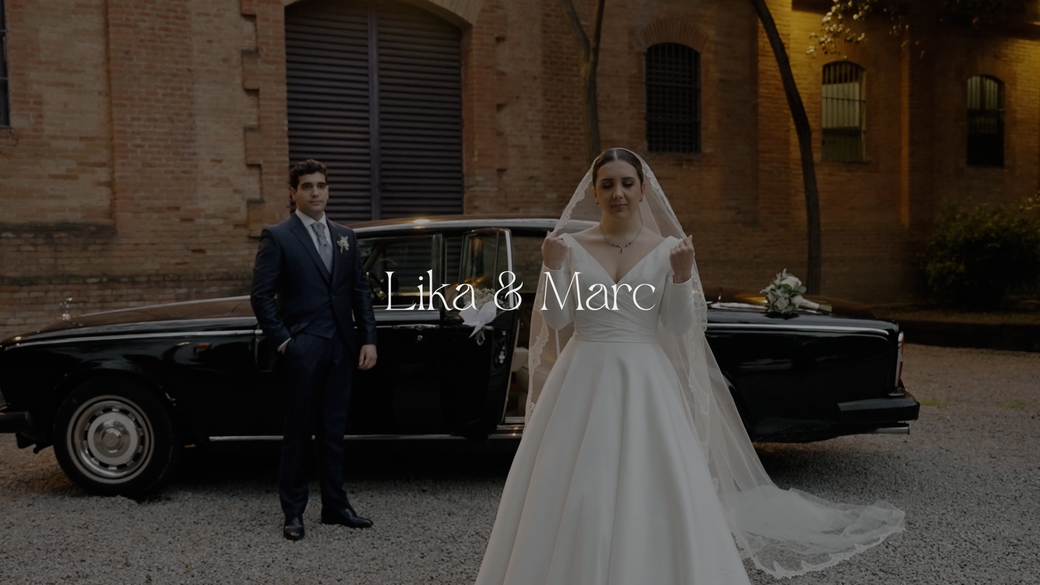 A couple on their wedding day pose beside a classic black car outside a historic brick building.