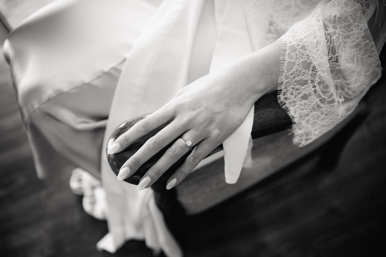 Black and white close-up of hands wearing delicate lace and a wedding ring against a dark background.
