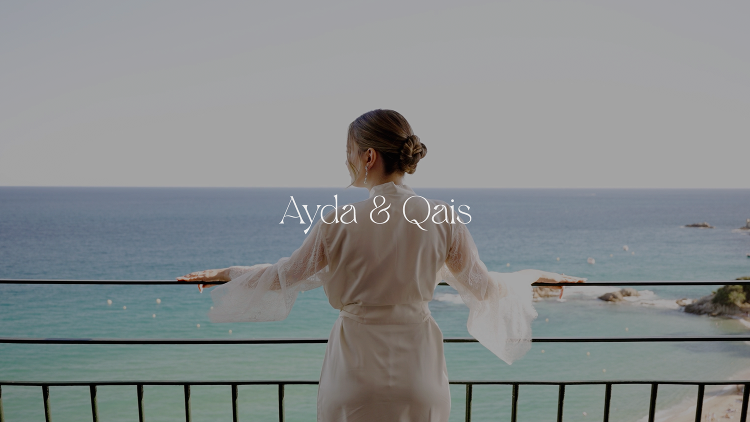 Person in white robe stands at seaside balcony overlooking blue Mediterranean waters on sunny day.