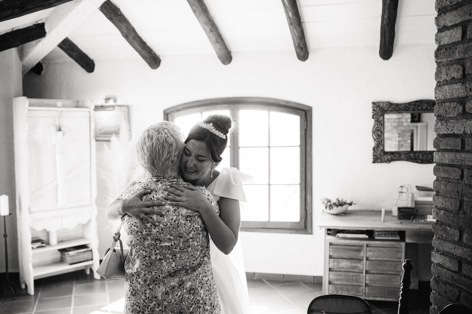 Black and white photo of two people embracing in a rustic room with wooden beams and arched windows.