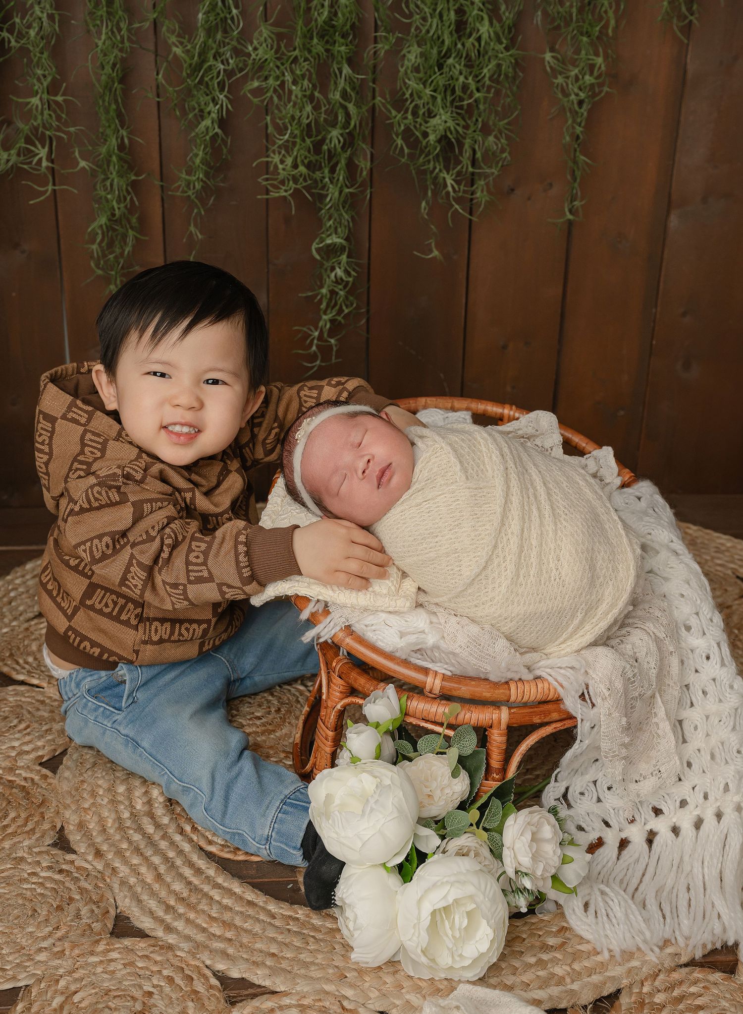 Two children sitting on bohemian-style textiles and decor featuring cream roses and wooden accents against a brown fence.