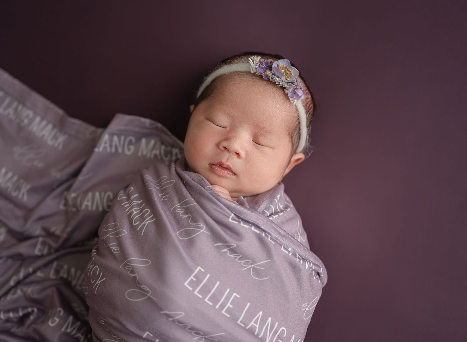 Sleeping newborn wrapped in soft purple blanket wearing floral headband during professional photo session.