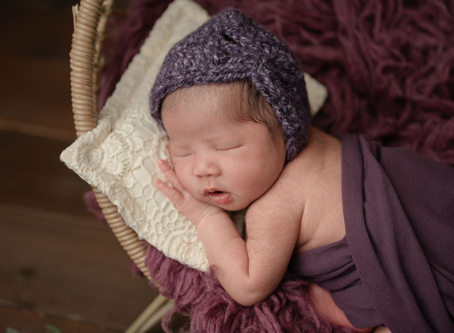 A sleeping newborn baby wearing a purple knit cap rests peacefully on cream and burgundy textiles.