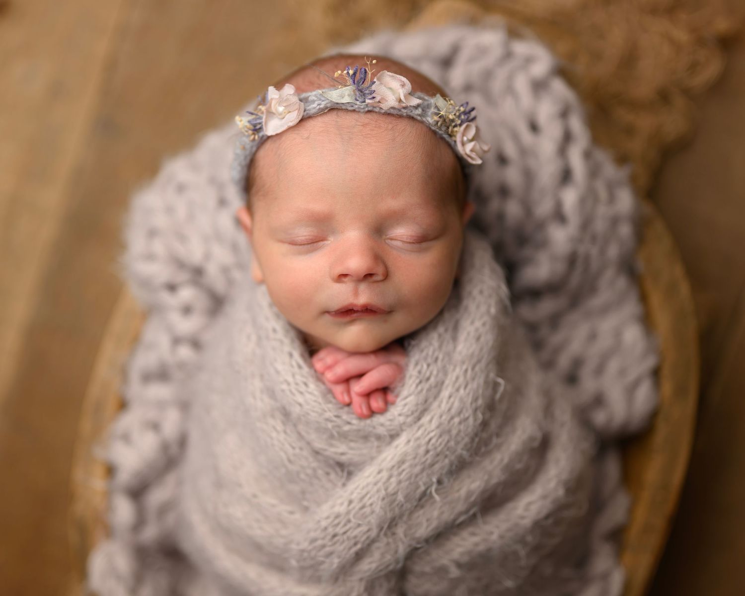 A sleeping newborn baby wrapped in soft gray swaddle blanket with delicate headband against brown backdrop.