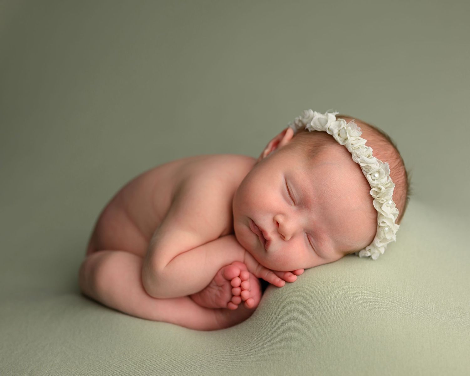Sleeping newborn baby wearing a delicate white headband curled up in peaceful slumber on soft neutral backdrop.
