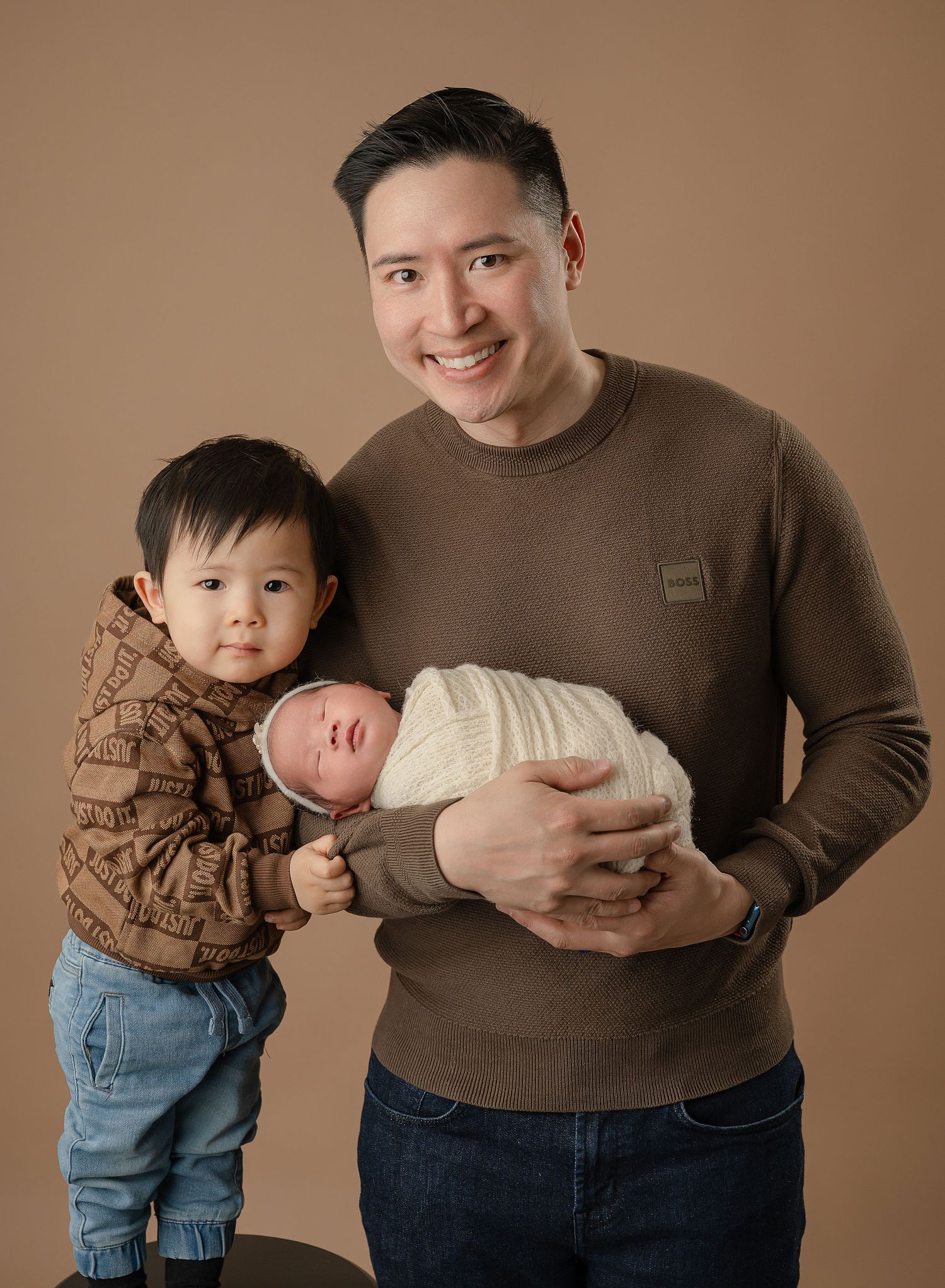 A heartwarming family portrait showing a parent holding a newborn baby while a toddler stands beside them against a brown background.