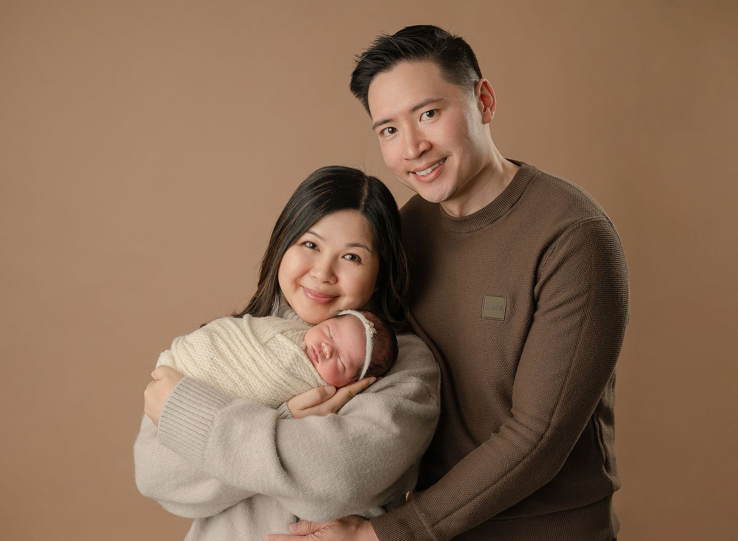 A family portrait featuring parents holding their newborn baby against a warm brown background.