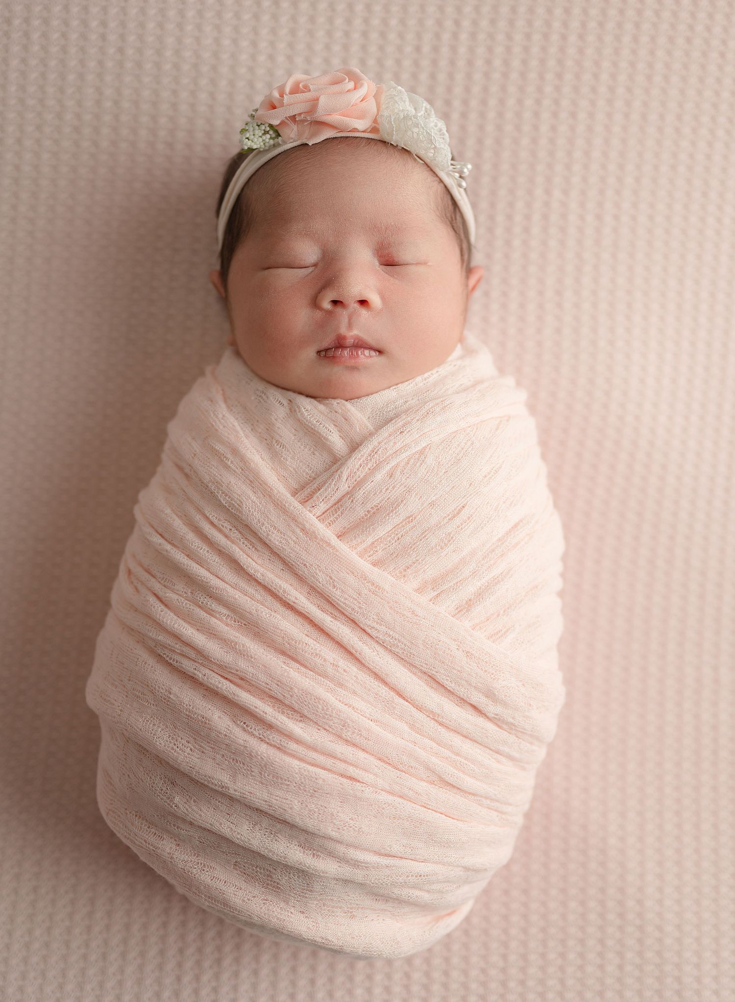 A sleeping newborn wrapped in soft pink cloth wearing a delicate floral headband against a blush background.