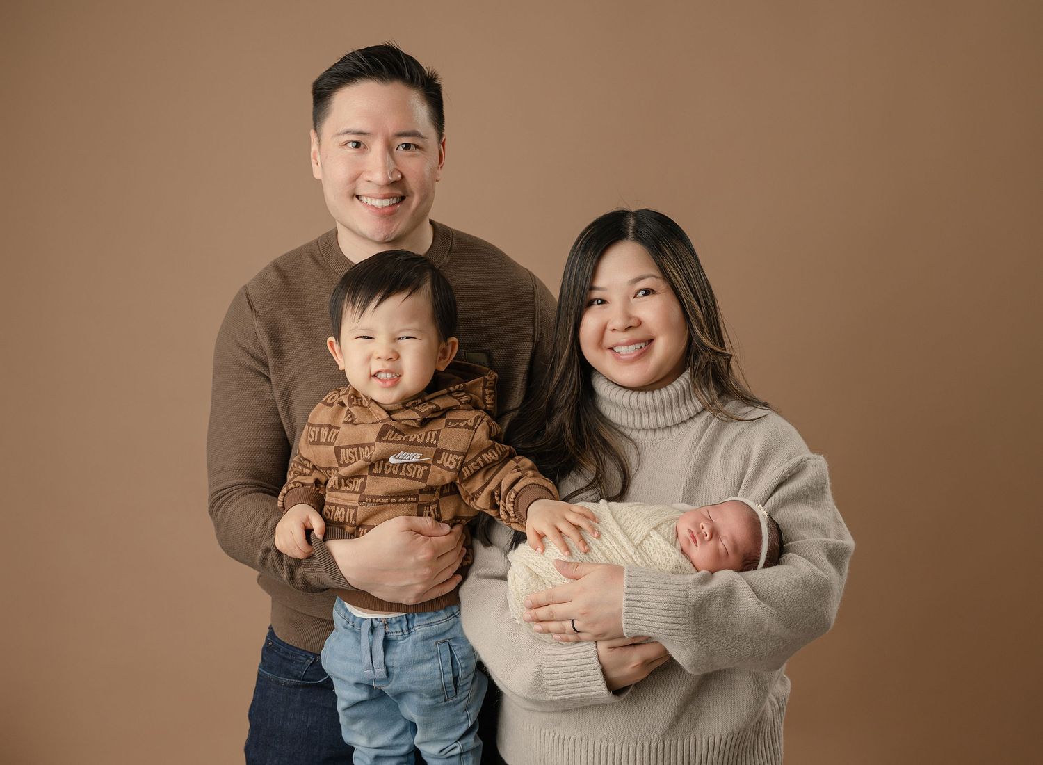 Family portrait against brown backdrop with parents wearing neutral sweaters and young children in casual attire.