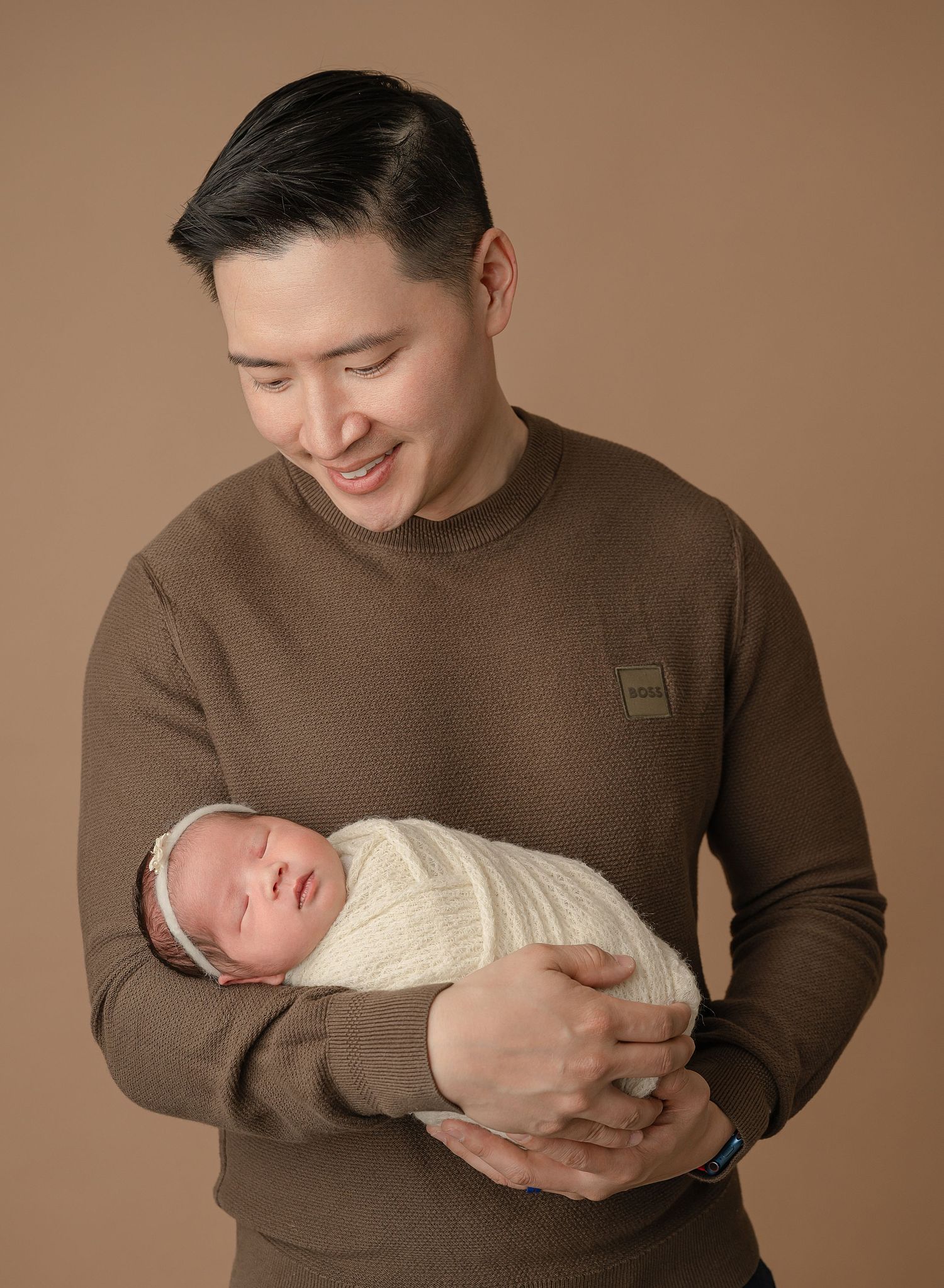 A person in a brown sweater tenderly holds a sleeping newborn baby wrapped in a cream-colored swaddle against a brown backdrop.