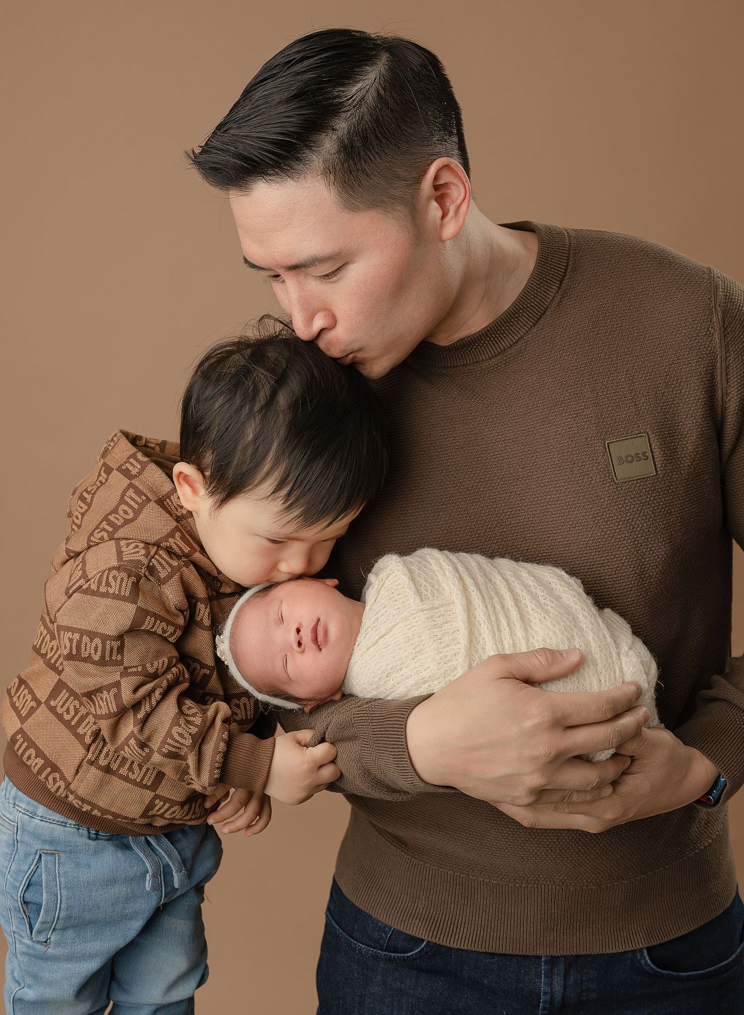 A tender family moment as a parent in a brown sweater embraces two children against a neutral brown background.