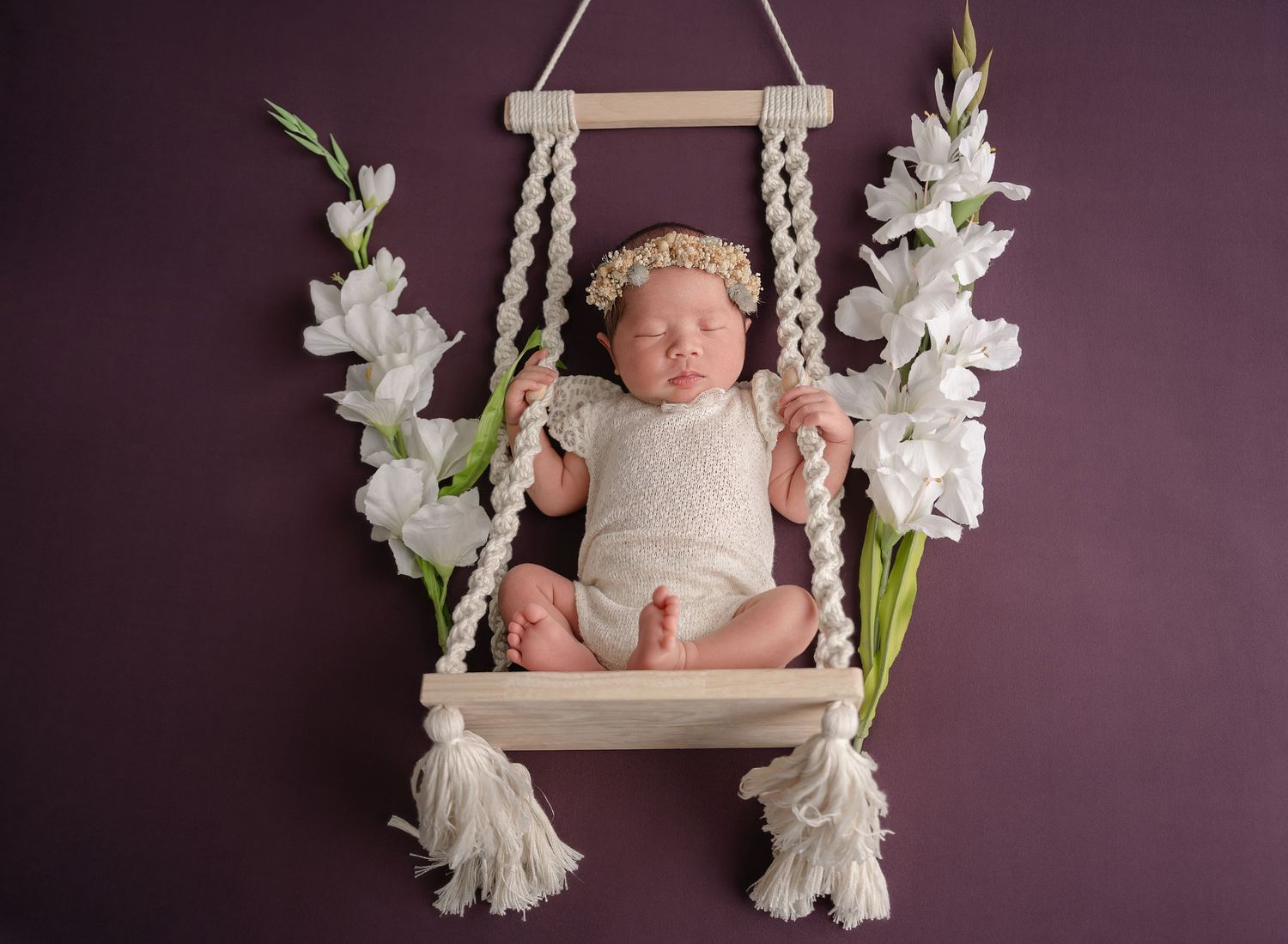 Sleeping newborn in white outfit rests on macrame swing decorated with white gladiolus flowers against purple wall.