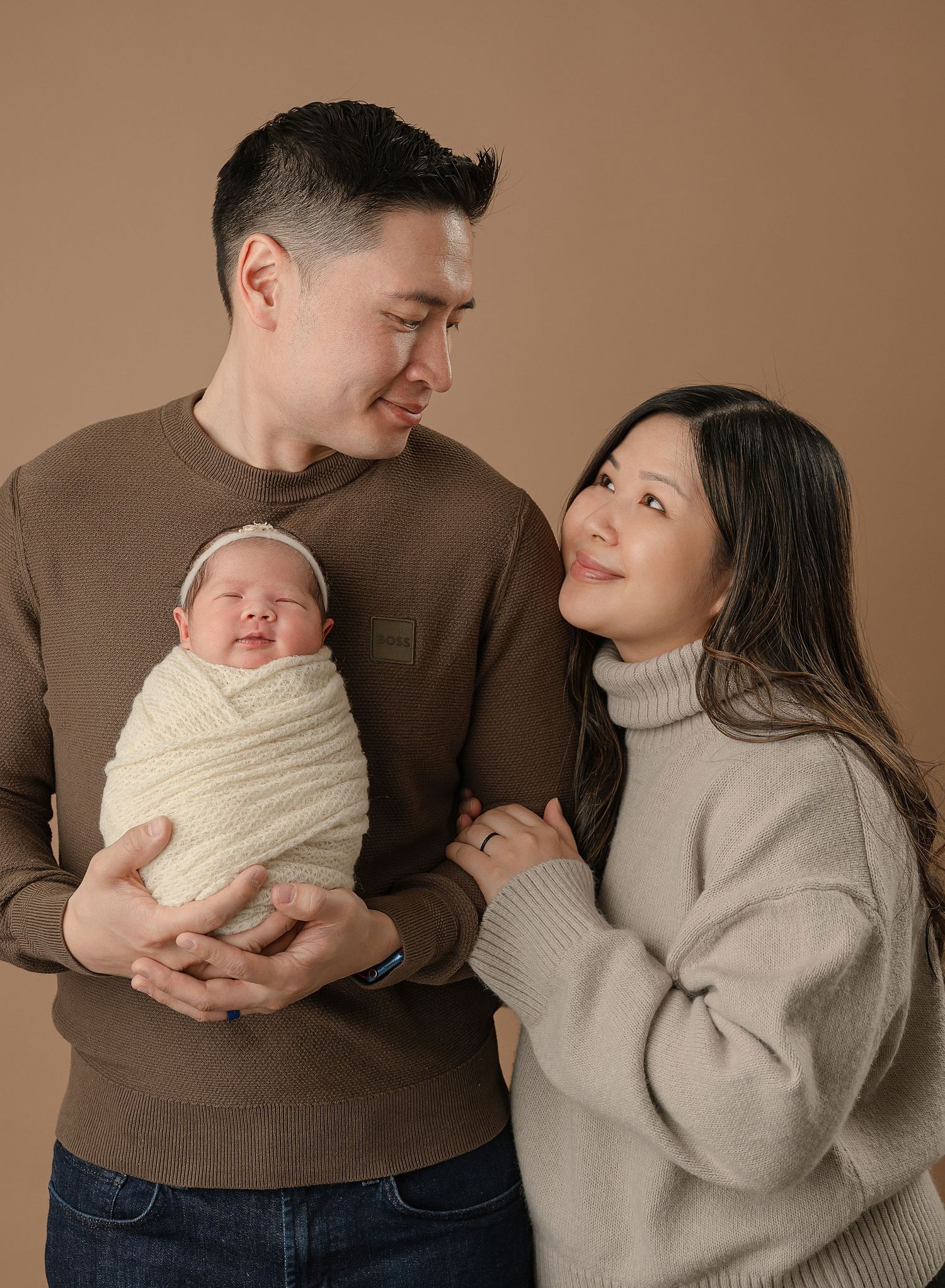 Parents in cozy sweaters share a tender moment with their sleeping newborn swaddled in cream-colored wrap against beige backdrop.