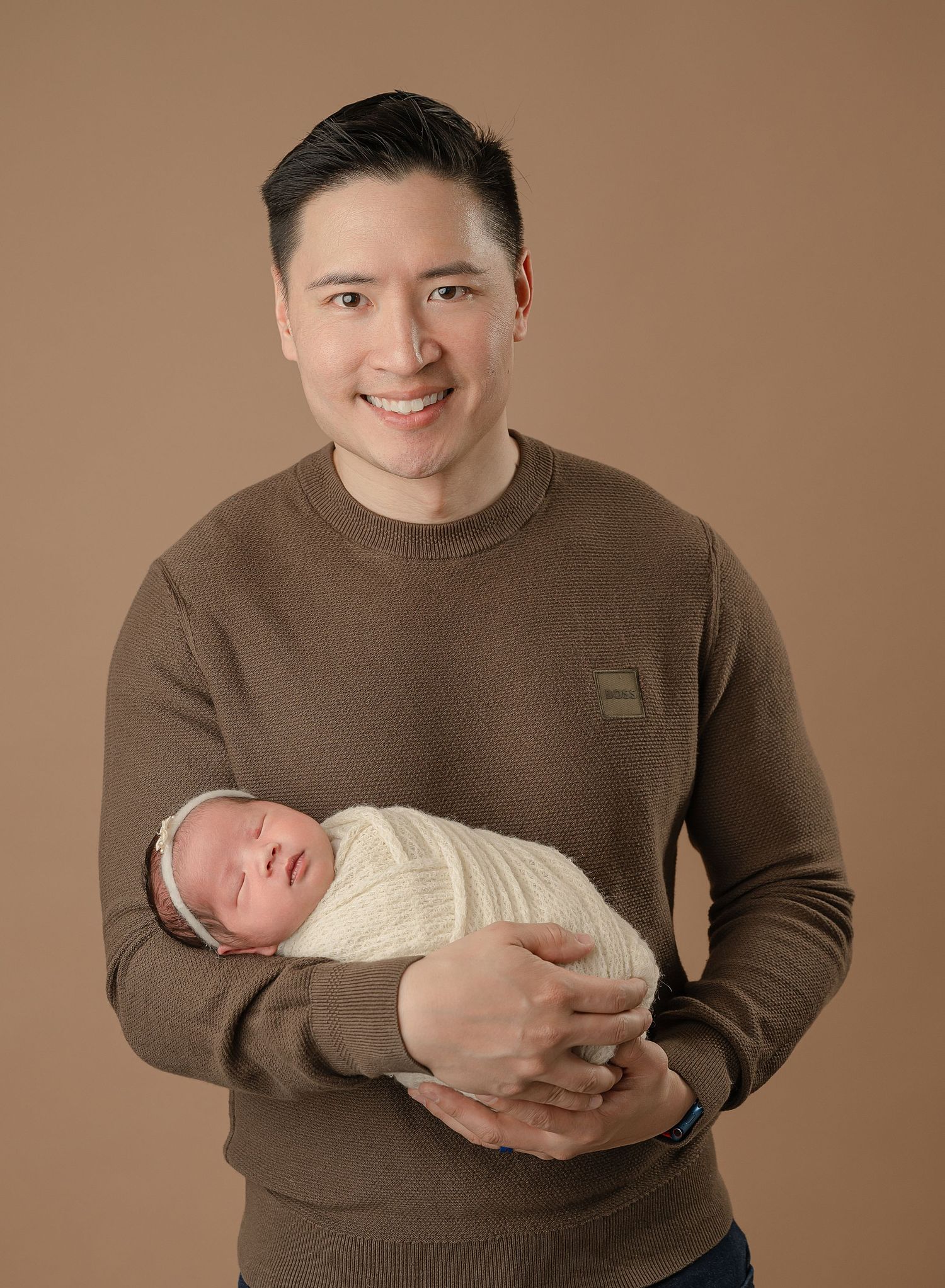 A tender moment of a parent in a brown sweater holding their newborn baby swaddled in white against a brown backdrop.