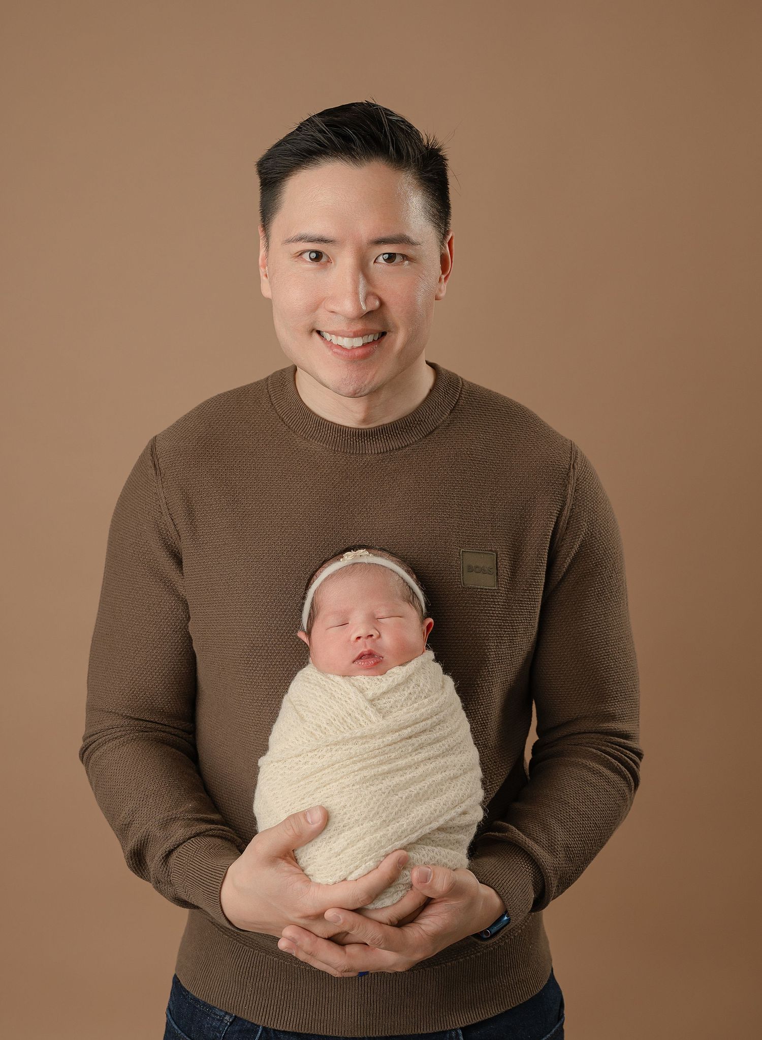 A tender portrait with a parent holding a swaddled sleeping newborn against a brown background.