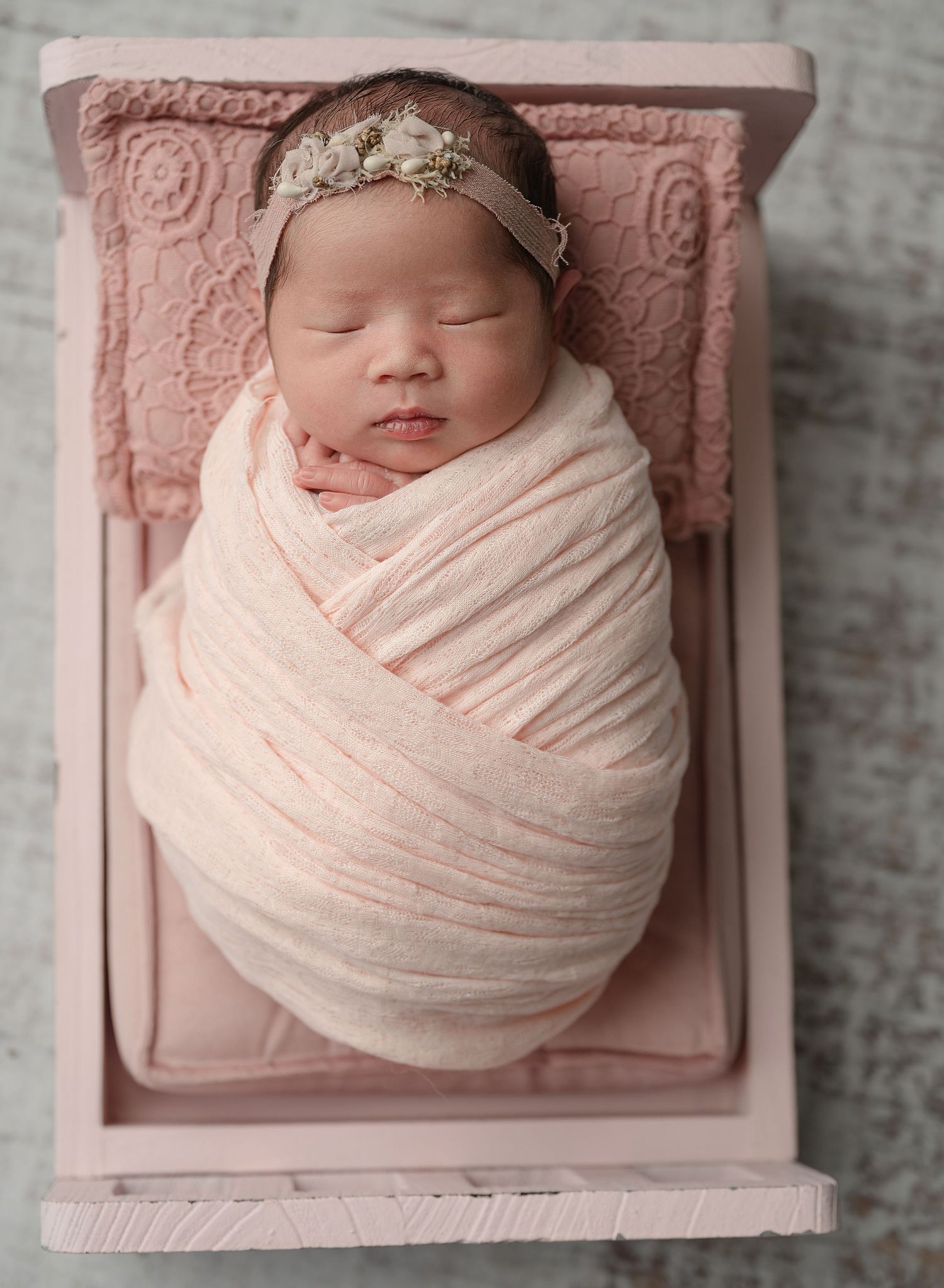 Sleeping newborn wrapped in soft blush pink swaddle rests in vintage pink wooden bed with delicate lace headpiece.