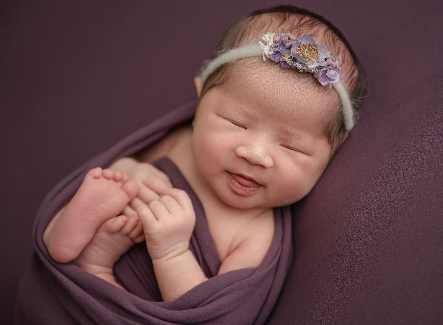 Sleeping newborn wearing floral headband wrapped in purple swaddle with hands clasped.