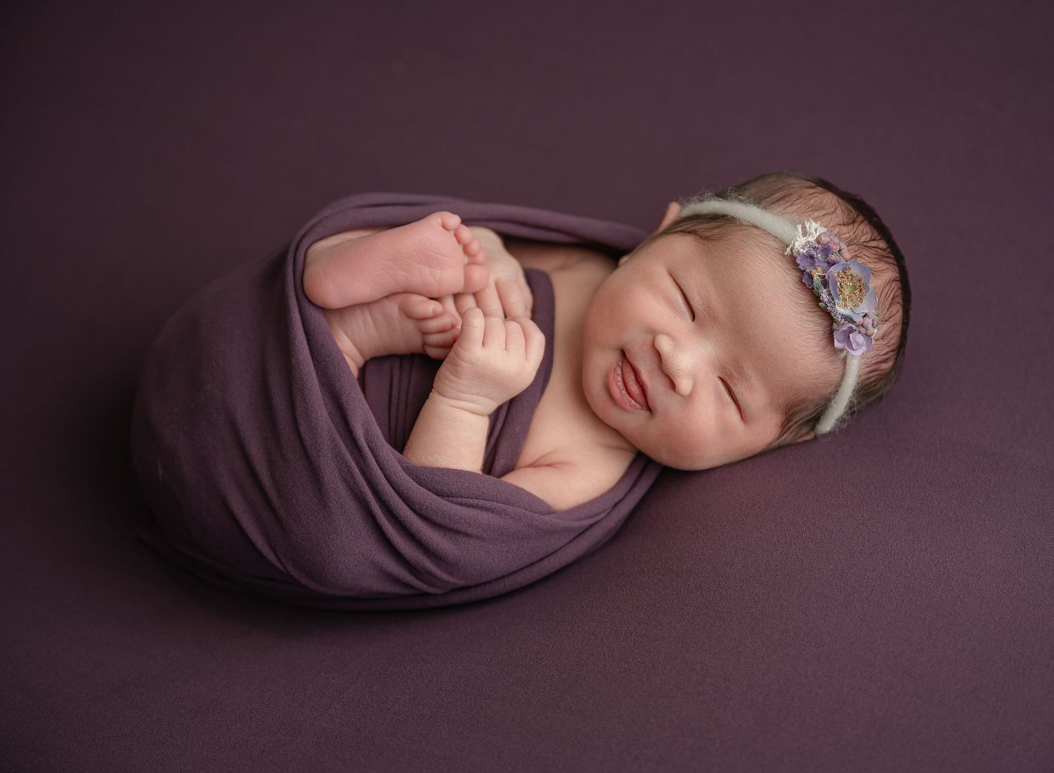 Newborn baby wrapped in purple swaddle cloth wearing floral headband sleeps peacefully during professional photoshoot.