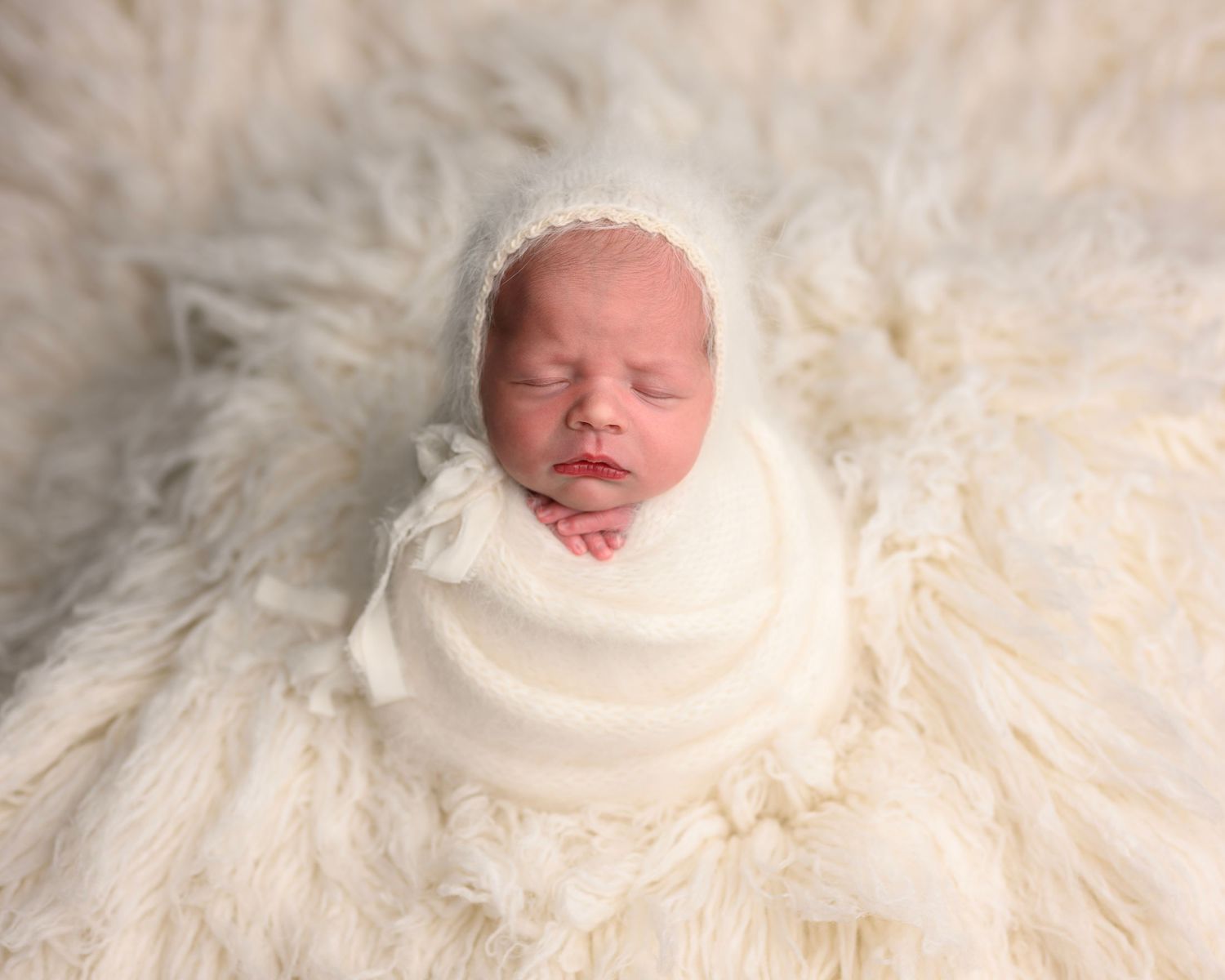 A sleeping newborn baby swaddled in white fabric lying on a fluffy white textured blanket.