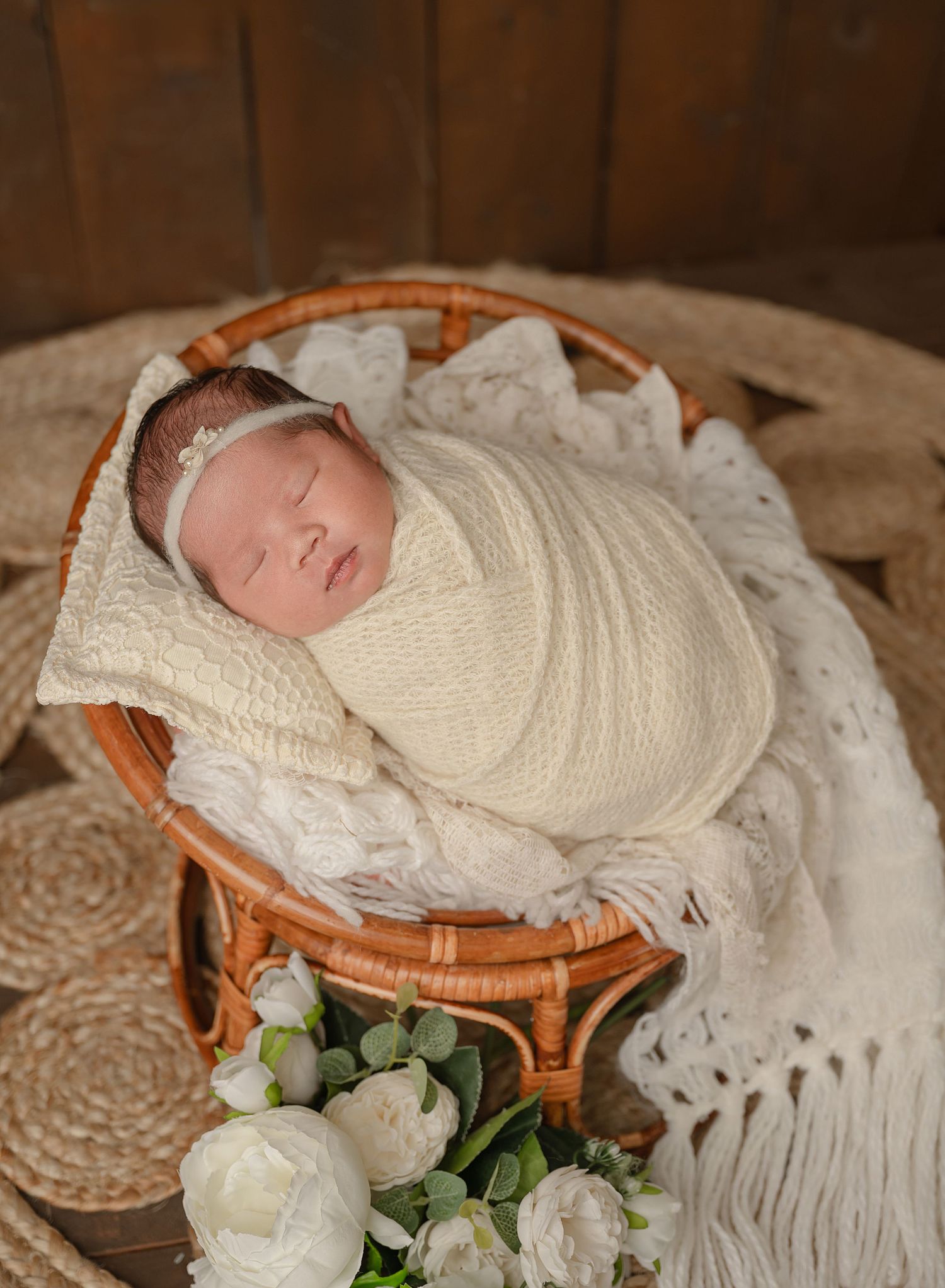 A sleeping newborn wrapped in cream-colored fabric rests in a wicker basket with white lace and floral decorations.