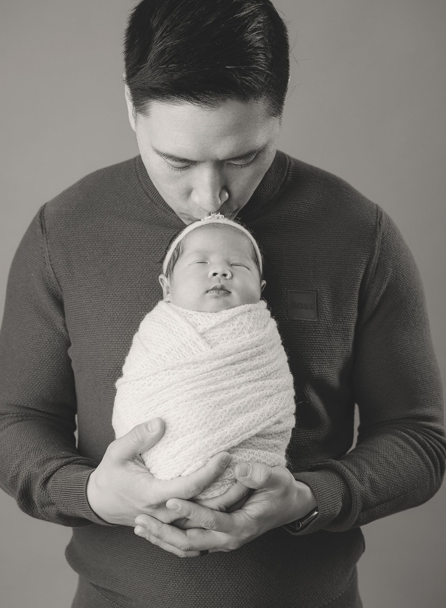 A tender monochrome portrait shows a parent cradling a swaddled newborn while giving a gentle kiss on the head.