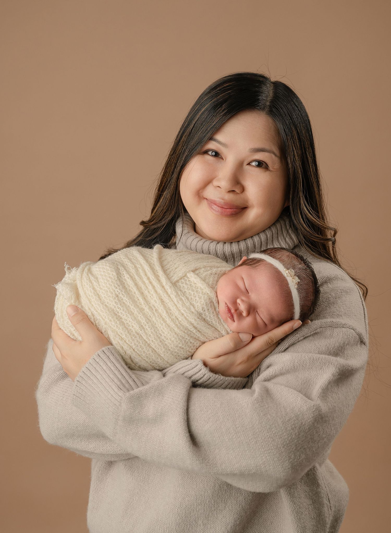 In a warm studio portrait against brown backdrop, parent in gray sweater cradles sleeping newborn wrapped in cream blanket.