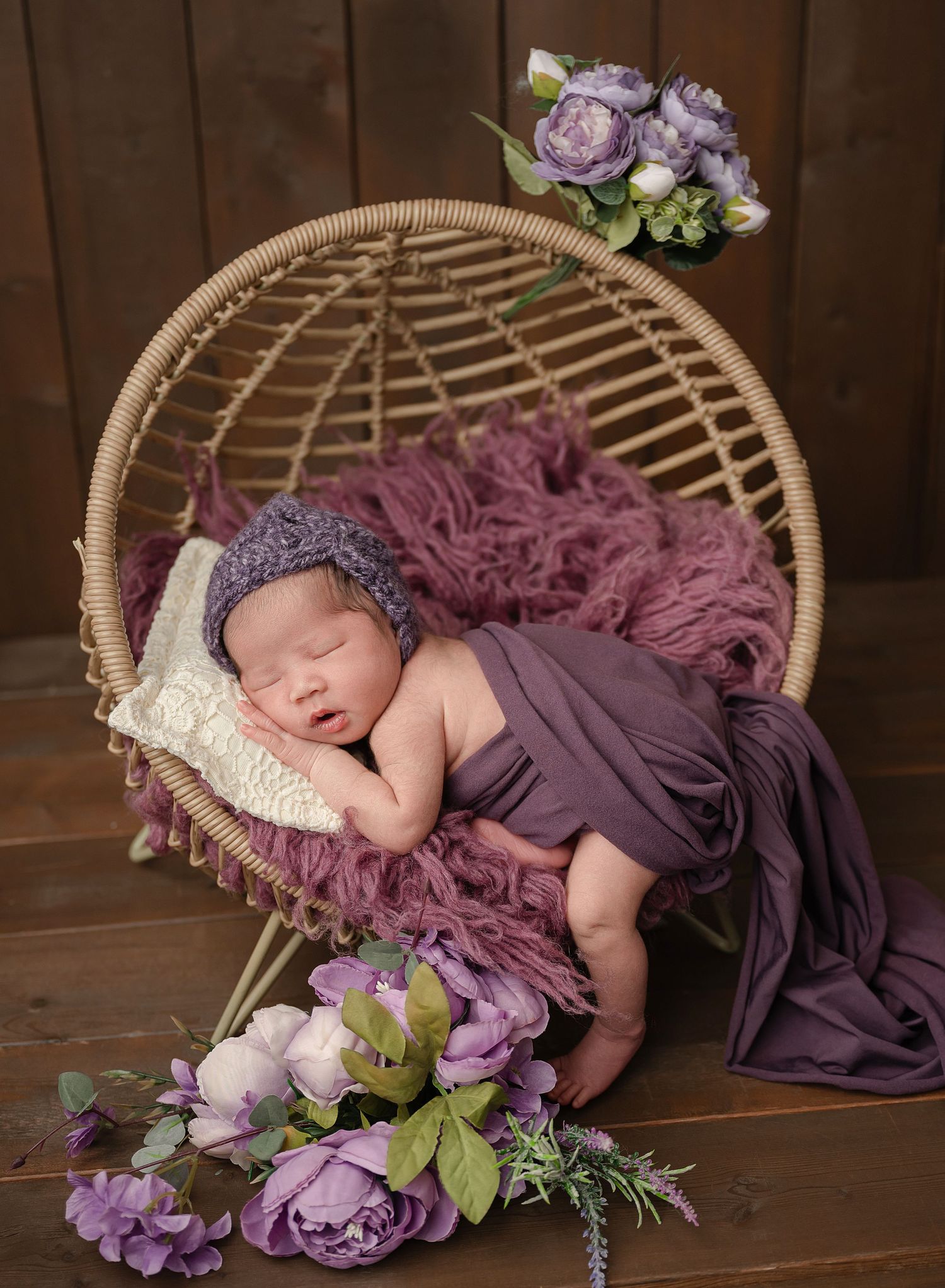 A newborn rests peacefully in a rattan chair with purple fabric and floral decorations in a rustic setting.